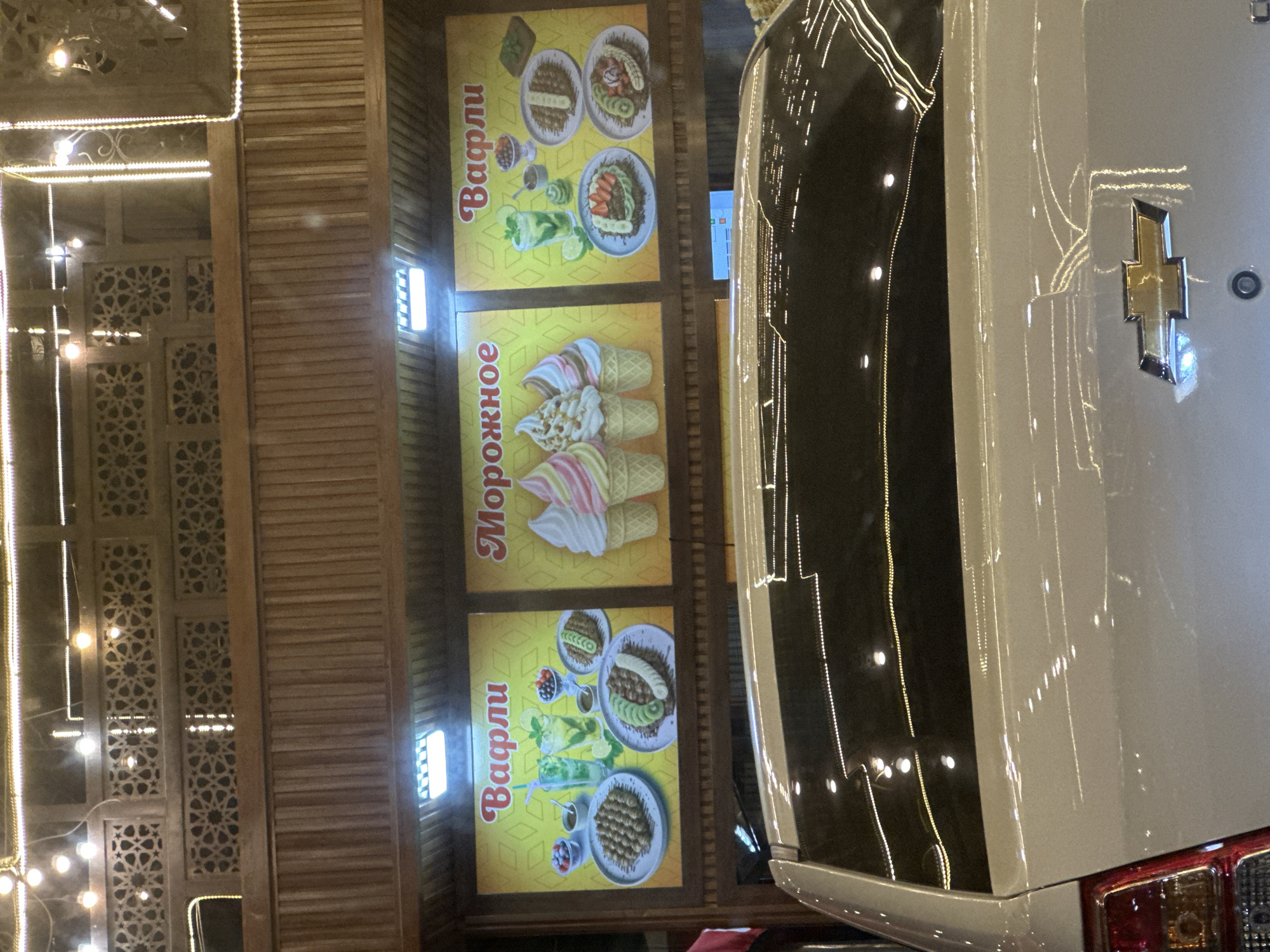 The image shows a nighttime outdoor scene in front of a food establishment. Three brightly lit signs are prominently displayed against a wooden facade. The signs are advertising "Вафли" (Waffles) on the left and right, and "Морожное" (Ice Cream) in the middle. Each sign features illustrations of the respective food items. The left "Waffles" sign shows several waffles, drinks, and berries. The "Ice Cream" sign displays three cones of swirled ice cream. The right "Waffles" sign also shows waffles, a drink, and berries. The establishment appears to have a decorative wooden exterior with intricate patterns and is illuminated by string lights. In the foreground, the rear of a white car is visible, reflecting the lights from the establishment. The image has a slightly grainy texture, suggesting it was taken with a mobile phone camera in low light conditions.