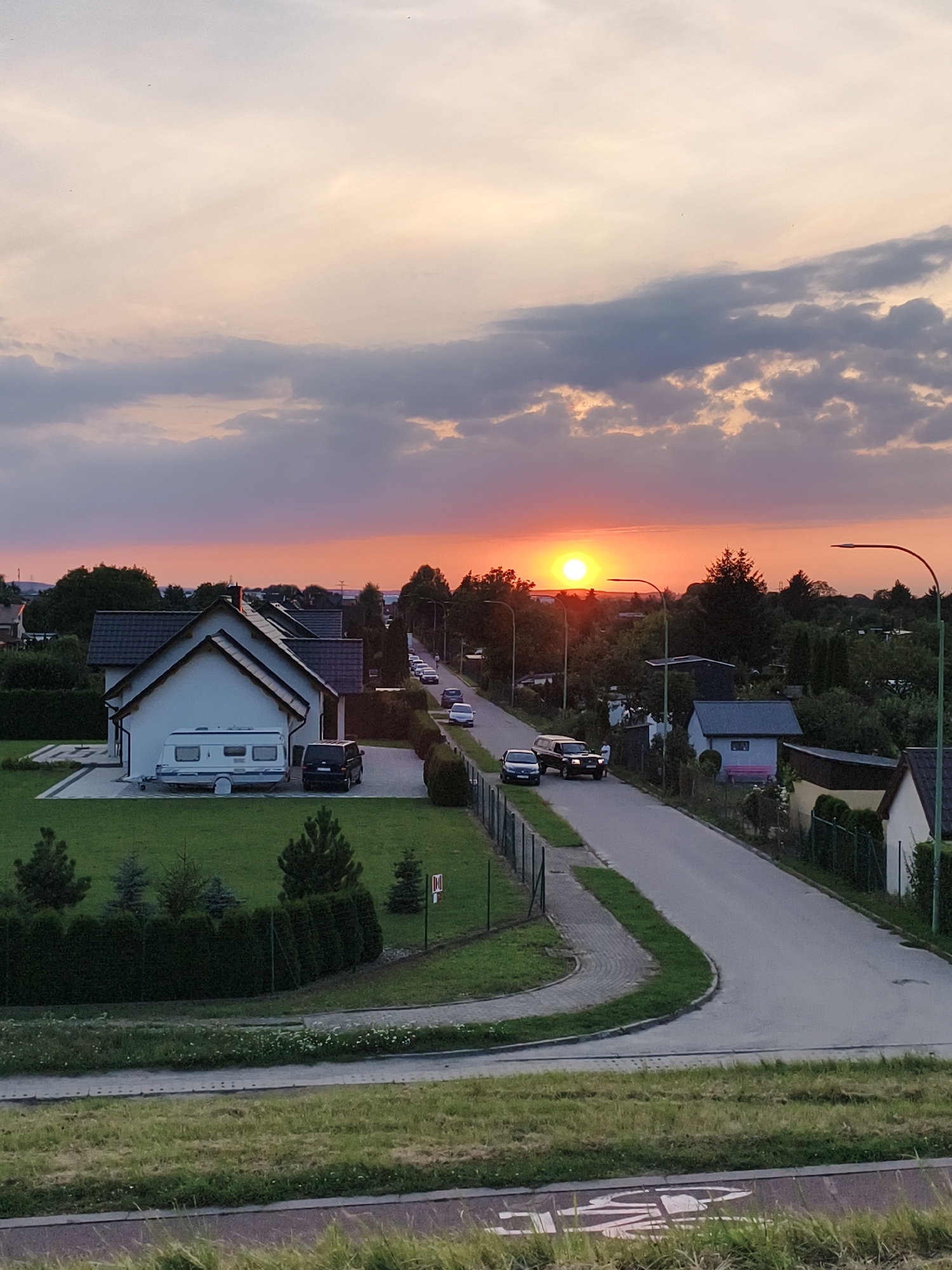 The image depicts a sunset over a suburban residential area in the city of Tczew, Poland. The sky is painted with warm hues of orange, pink, and yellow, with scattered gray clouds. In the foreground, a grassy embankment slopes down towards a paved path. Beyond the path, a well-maintained lawn leads to a white house with a dark roof, a caravan parked in the driveway, and a black van. A road curves to the right, lined with houses, fences, trees, and streetlights. Several cars are visible on the road, including a black SUV. The overall scene suggests a peaceful evening in a residential neighborhood.