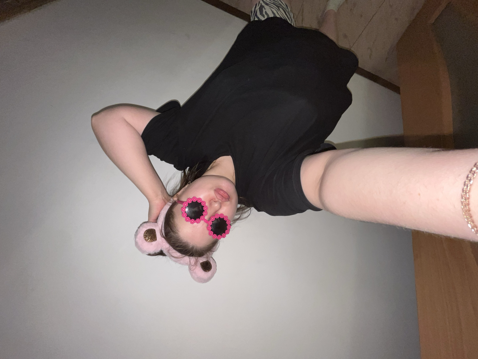 A person wearing a black t-shirt, pink bear ears headband, and pink flower-shaped sunglasses is taking a selfie. The background includes a white wall and a portion of a wooden door or furniture. There is a piece of zebra-striped fabric visible in the top part of the image, possibly indicating bedding or a cushion. The location is an indoor space.