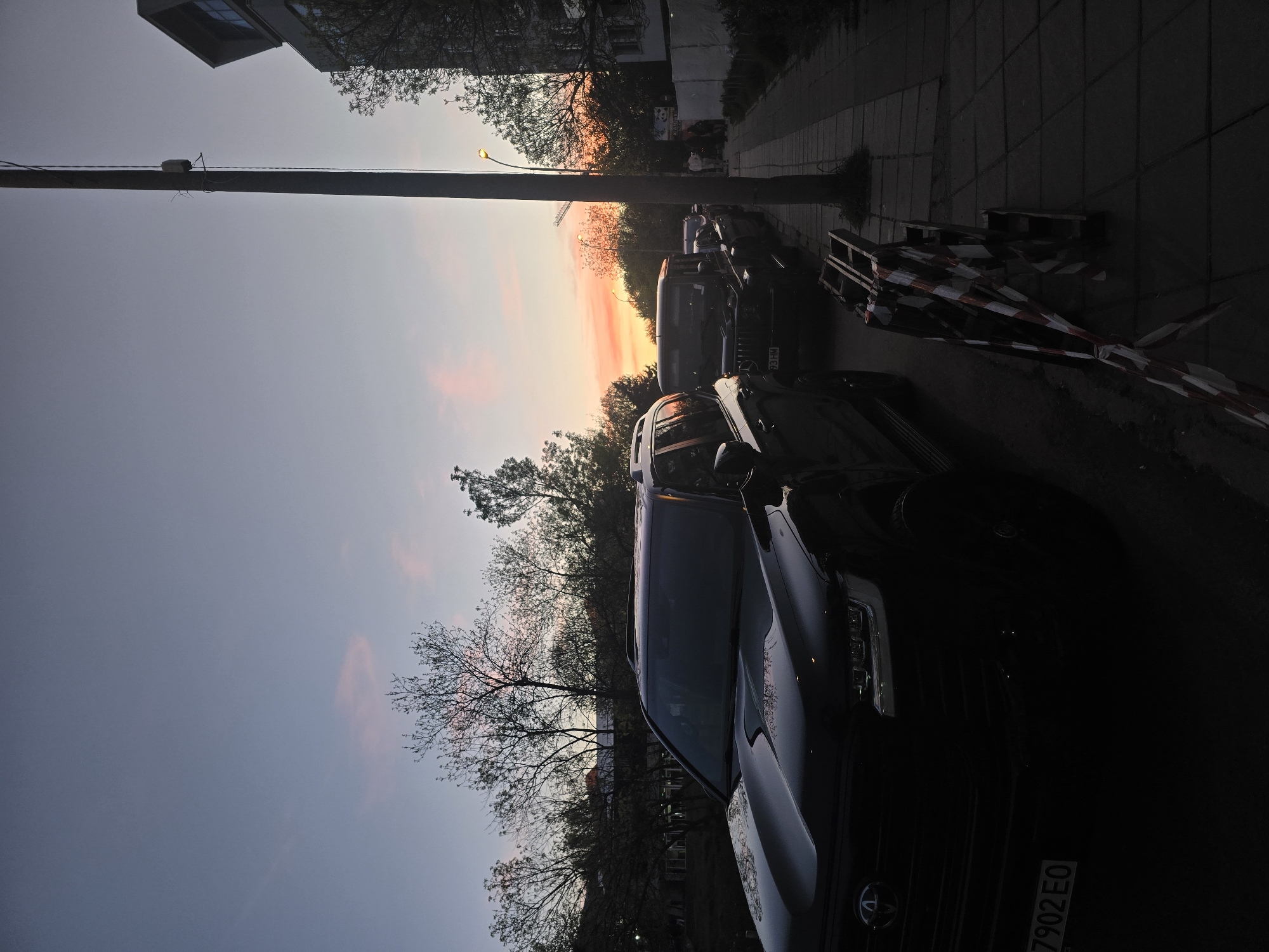 The image displays an urban street scene in Sofia, Bulgaria, captured during sunset or dusk. The sky dominates the upper portion of the frame, exhibiting a stunning gradient of colors: a soft, light blue-grey at the top transitions into vivid orange, pink, and yellow hues closer to the horizon, indicative of the setting sun.
Silhouettes of numerous deciduous trees, mostly bare or with sparse leaves, are visible against the colorful sky, creating a stark contrast. The tops of buildings appear as dark shapes in the distance, further contributing to the urban setting.
A prominent, dark utility pole runs through the left side of the image, with some small equipment attached. Along the right side of the street, several cars are parked, mostly appearing as dark masses in the diminishing light. In the immediate foreground on the right, the front section of a dark SUV, likely a Toyota given the visible logo, is partially visible. Its license plate clearly shows the partial text "7902 EO." Further down the street, another dark vehicle is parked, and a streetlight glows with a warm yellow light. A paved sidewalk runs parallel to the street, and in the mid-ground on the right, a temporary barrier marked with red and white striped tape suggests a construction or restricted area.
No specific human activity is discernible, lending a tranquil and contemplative mood to the scene as day transitions into night over a quiet city street. The weather appears clear, with no clouds obscuring the colorful sunset.