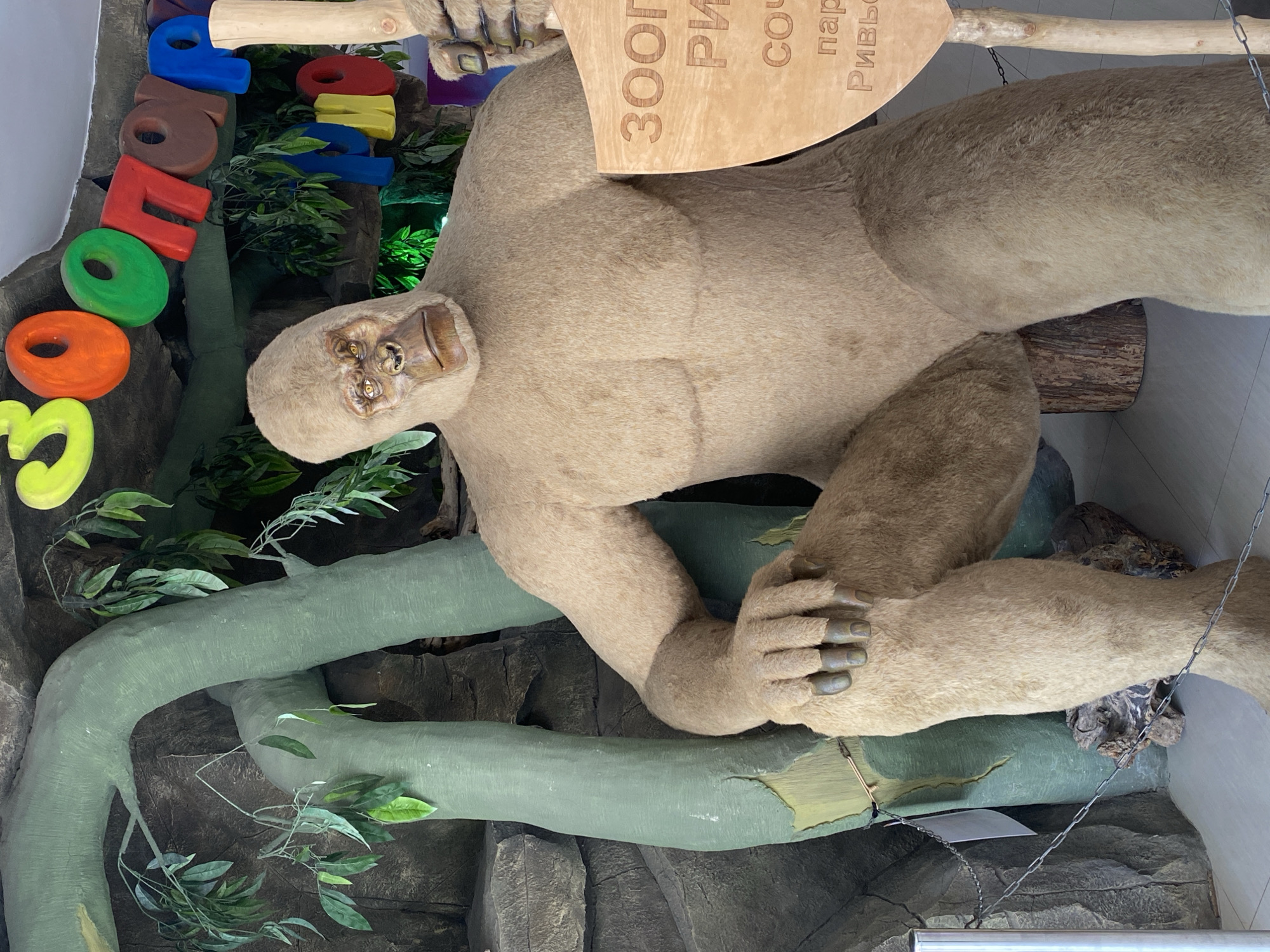 The image displays an indoor exhibit featuring a large, inanimate figure resembling a light-brown, furry primate or sloth, suspended horizontally. The figure has a stylized face with yellow eyes and a prominent snout. It rests its body on artificial green tree branches and textured rock formations, with one arm extended to grasp a wooden pole. Its hands and feet have defined digits with darker nails.
The setting appears to be a zoo or park exhibit, consistent with the provided location context of Sochi, Russia. Artificial foliage and tree-like structures create an immersive, jungle-themed environment. The scene is well-lit by interior lighting.
Visible on the left side of the frame are colorful, three-dimensional block letters spelling out the Russian word "ЗООПАРК" (ZOOPARK). Above the figure's body, a carved wooden sign is partially visible, with legible Russian text fragments that likely combine to form "ЗООПАРК РИВЬЕРА" (ZOOPARK RIVIERA), or "ЗООПАРК СОЧИ РИВЬЕРА", indicating a specific location within the Riviera park in Sochi. The overall scene depicts a static display designed for public viewing at a recreational or educational facility.