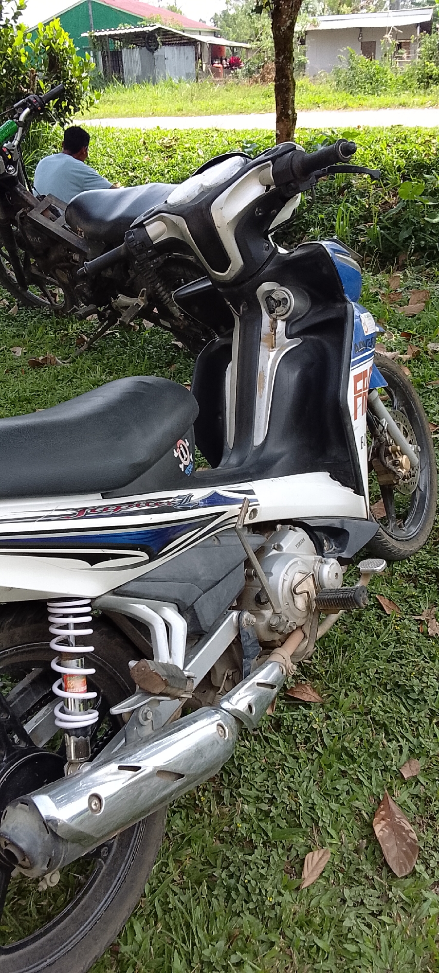 This is a ground-level, eye-level, vertical shot of two motorcycles parked on a grassy area. In the foreground, a white and blue motorcycle is parked with its front wheel slightly turned to the left. The motorcycle has a black seat and visible engine components, including the exhaust pipe. A person wearing a light blue shirt is seated on the rear of the other motorcycle, which is mostly obscured by the foreground motorcycle. The person is facing away from the camera. In the background, there are several small buildings, some with corrugated metal roofs, and trees. The environment appears to be rural, consistent with the location of Padang Dalom, Indonesia.