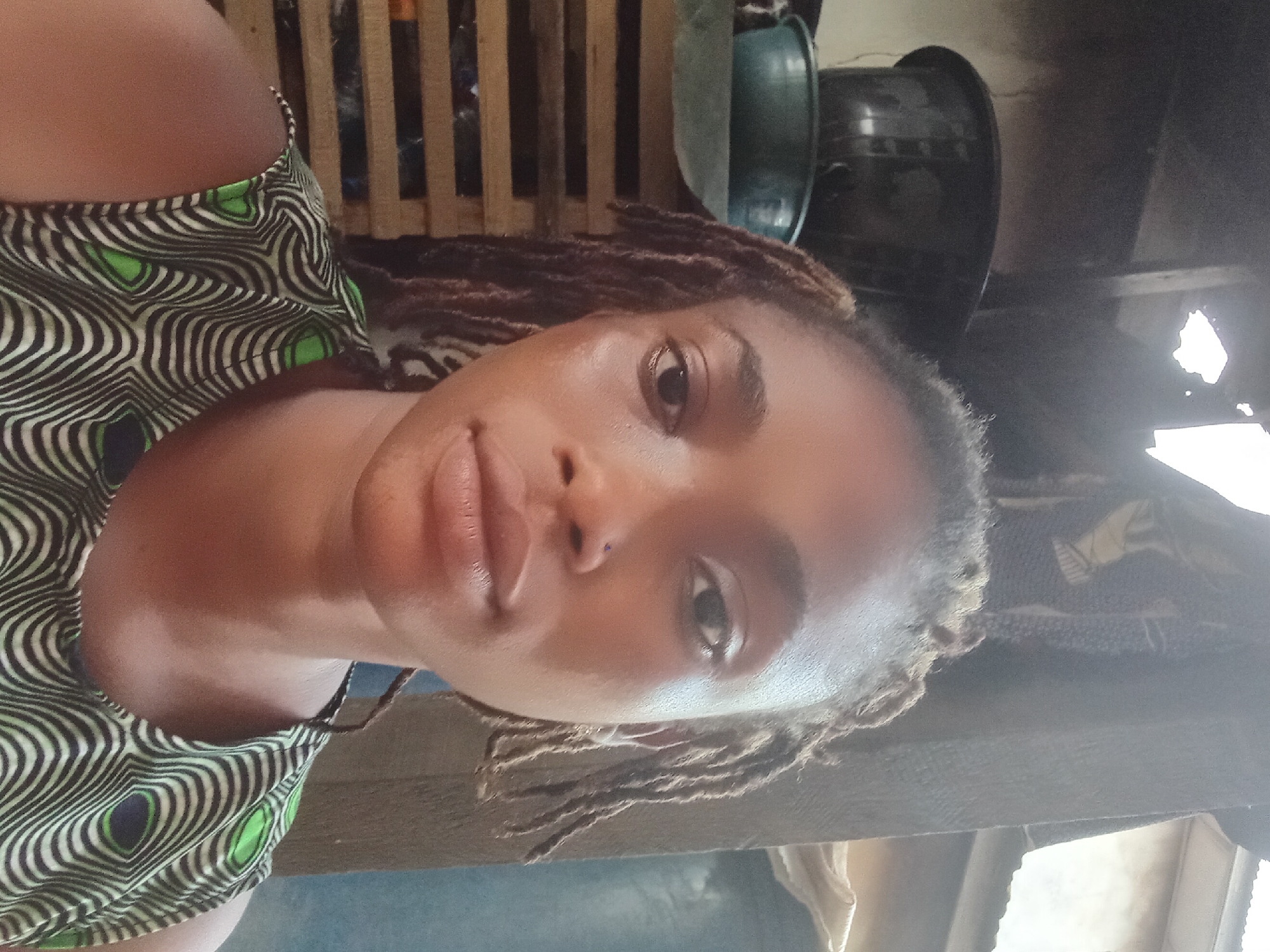 This close-up indoor selfie features a young woman with medium-brown skin and dark brown dreadlocks, captured in Shagamu, Nigeria. The image is rotated 90 degrees counter-clockwise, showing her face in a slight upward gaze with a neutral to subtly pleasant expression. She is wearing a sleeveless top adorned with a striking pattern of wavy black lines against a light background, accented by circular elements in bright green and dark blue/black.
The background on the left side reveals a wooden slatted structure, possibly part of a wall or furniture. To the right of the woman's head, two dark, utilitarian containers—one green and one black bowl—are visible. Further right, a bright, irregular patch of light suggests a window or opening, indicating natural daylight illuminating the scene. No discernible activities other than posing for the selfie are taking place. The time of day appears to be daytime, given the natural light. There is no visible text in the image.