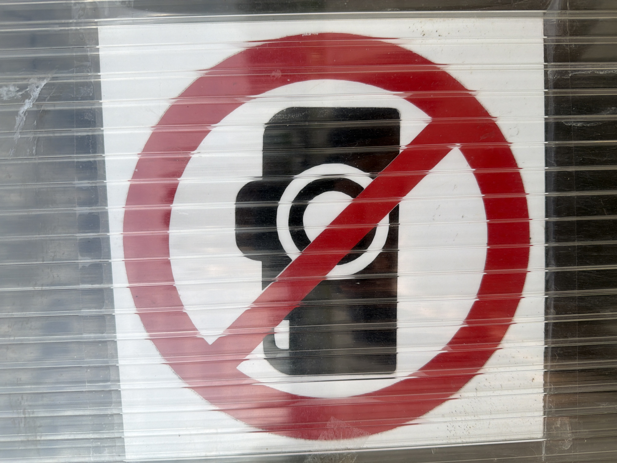 A photograph taken in the city of Sofia, Bulgaria, presents a "no photography" sign viewed through a translucent, vertically ribbed plastic panel. The sign features a black camera icon on a white rectangular background, overlaid with a standard prohibition symbol: a red circular border with a diagonal red line striking through the camera. The ribbed plastic material creates parallel vertical lines across the entire image, subtly distorting the visual elements of the sign and its surroundings. The plastic surface exhibits minor grime, smudges, and scratches. Above the white portion of the sign, a darker, mottled background is visible through the plastic. Below the sign, the plastic covers a dark, uniform surface. The red paint of the prohibition symbol appears slightly faded and worn in areas.