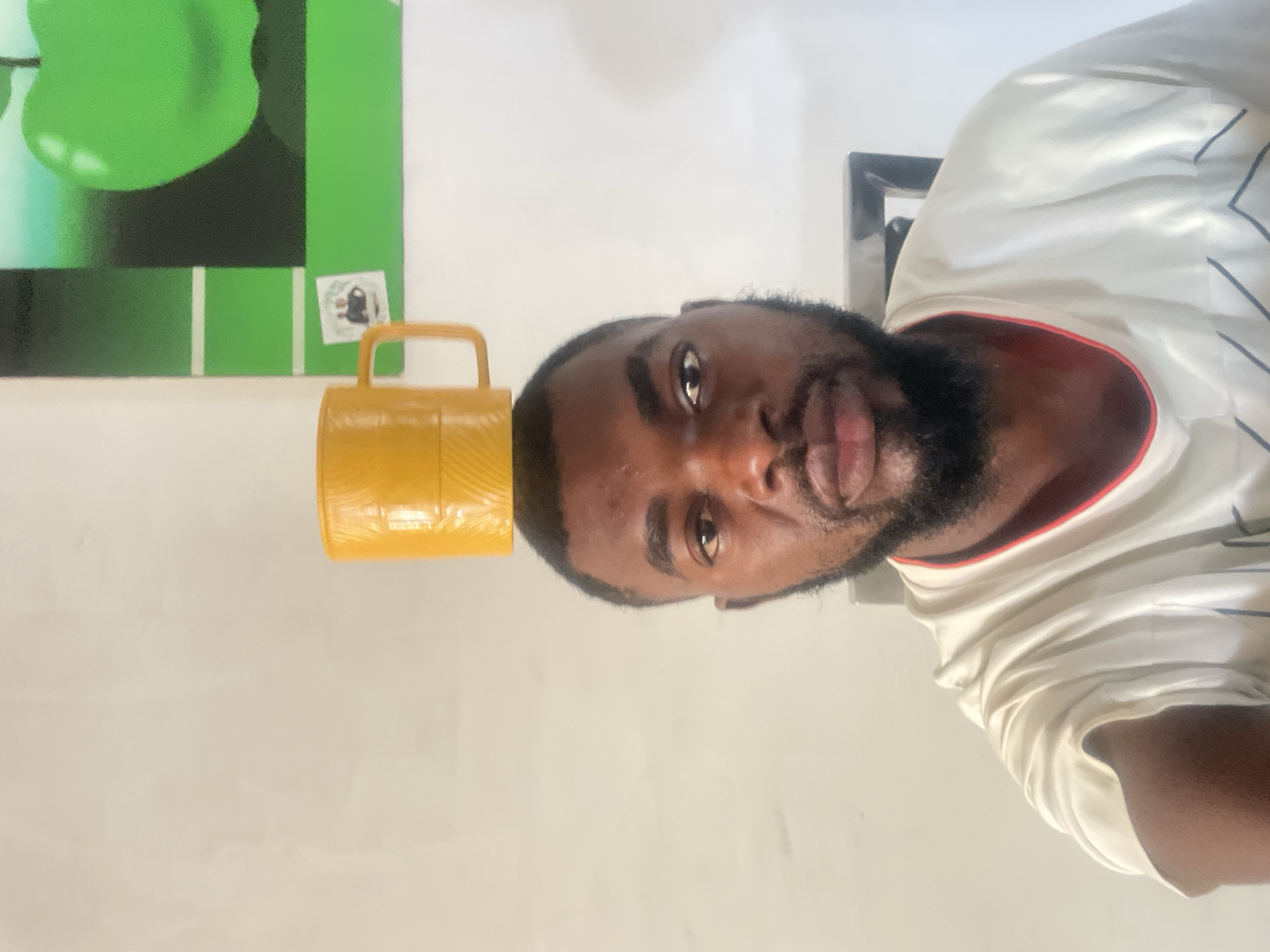 A close-up indoor shot shows a Black male with short hair and a beard looking directly at the camera, rotated clockwise. A yellow, plastic mug with a handle rests on top of his head. He is wearing a white t-shirt with red and navy accents. Behind him, on the left, is a wall with a green painting, and a white wall in the center. The location is inferred as Jos, Nigeria, based on contextual information not present in the image.