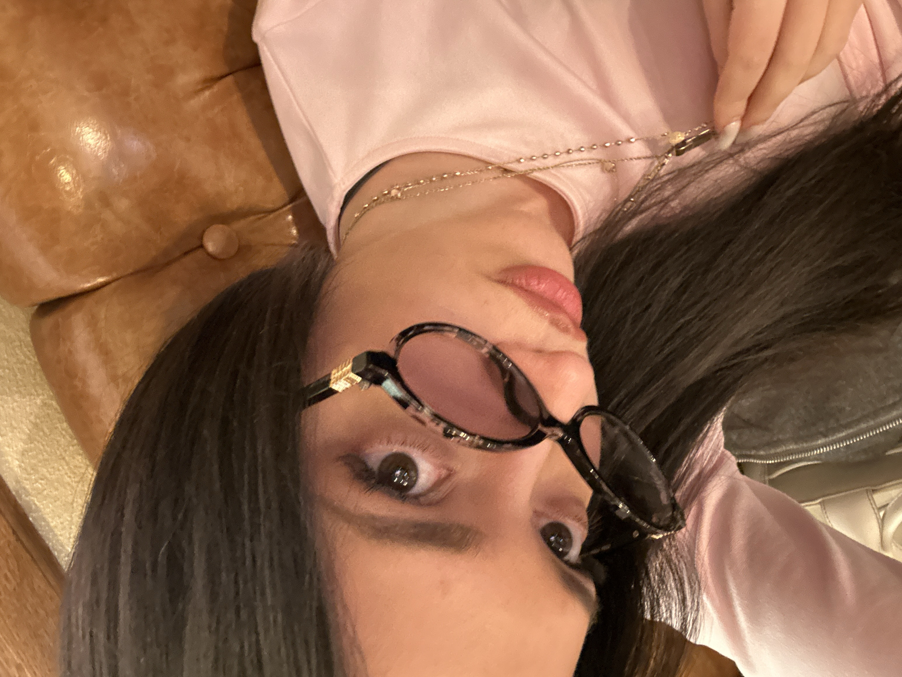 A woman lies on a brown leather couch, her head tilted back. She wears a pink shirt and a gold necklace. Dark hair frames her face. She sports sunglasses with patterned rims and pink lenses. One hand is visible, holding the necklace. The couch's texture and button detail are visible.