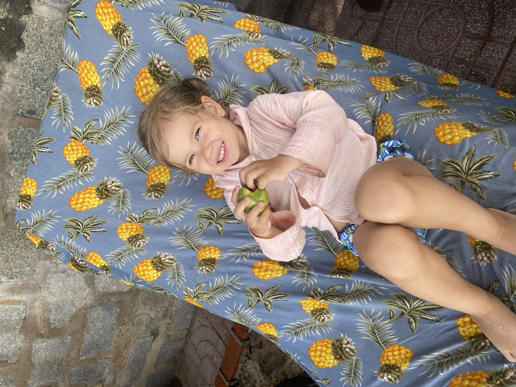 A young child with blonde hair tied in a bun lies on their back on a blue fabric with a pineapple pattern. They are wearing a pink, gauze-like shirt and patterned shorts. The child is smiling broadly at the camera, with their eyes open and looking directly at the viewer. They are holding a green object, possibly a fruit, in their right hand. The child's legs are bent, and their body is slightly angled. The fabric they are lying on is spread over an area that appears to be outdoors, with glimpses of stone and brickwork suggesting a patio or paved area. The lighting suggests it is daytime. The overall impression is one of a happy, relaxed moment.