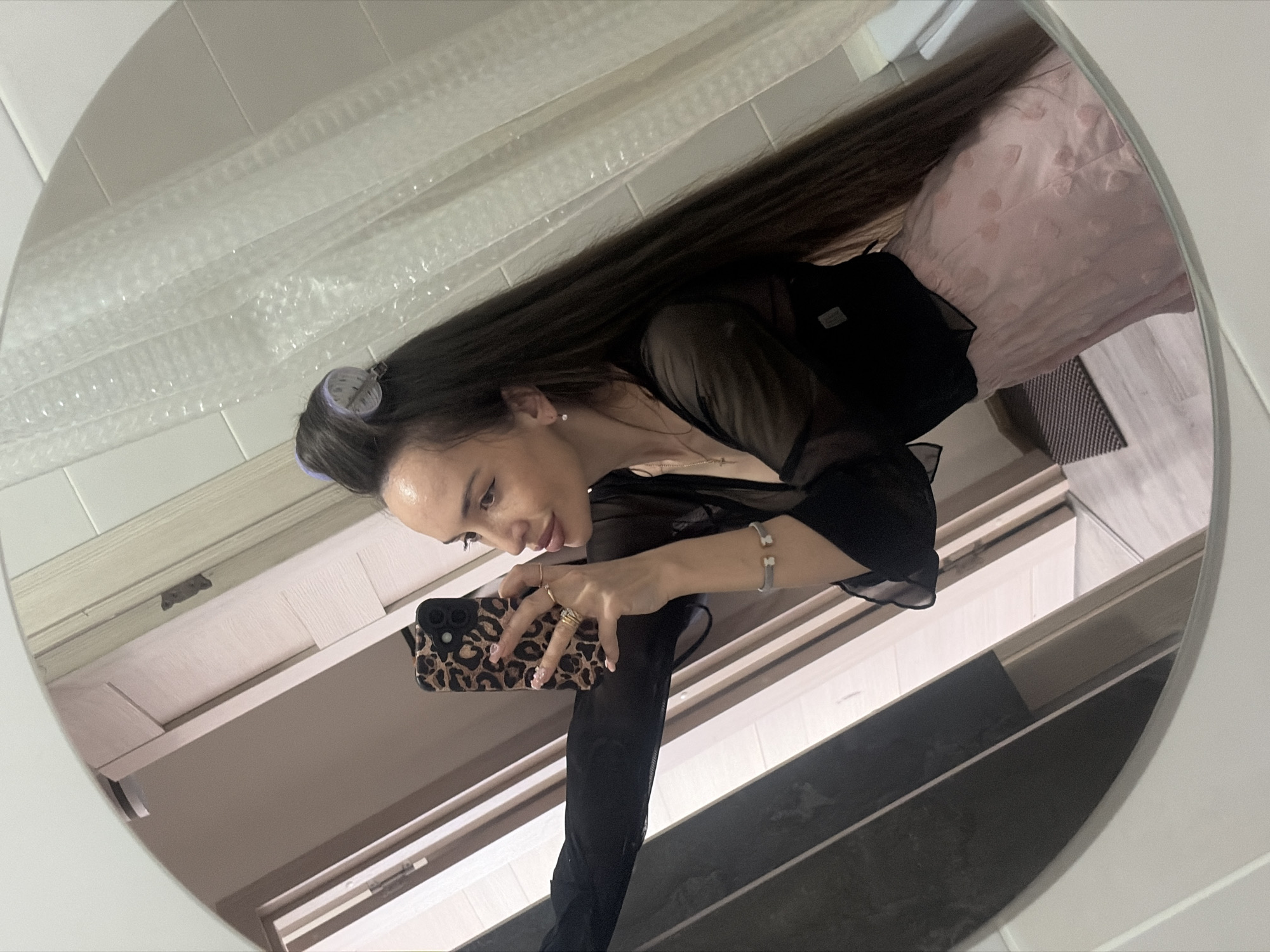 A woman is depicted taking a mirror selfie in what appears to be a bathroom, though the specific location in the city of Saint Petersburg, Russia, cannot be visually confirmed from the image. The subject is a woman with long, dark hair, wearing two purple hair rollers at the top of her head. She is clad in a sheer black top layered over a dark undergarment and a light pink skirt or shorts with a textured, possibly embroidered, pattern. Her right hand, adorned with multiple gold-toned rings and light pink nails with a white spotted design, holds a black smartphone encased in a leopard print cover. Her left wrist displays a silver-toned bracelet featuring two bear motifs. A delicate necklace with a small pendant and a single clear stud earring are also visible. She gazes towards the phone's reflection in the mirror, which captures a partial view of her face with visible eyeliner and plumped lips.
The mirror's reflection reveals the immediate environment: to the right of the woman, a white, translucent shower curtain with a bubble-like texture hangs against a tiled wall. To her left, a light-colored wooden door frame and a section of a darker wall or door are visible. The lower portion of the reflection shows a light-toned floor, possibly wood or laminate, with a dark, textured rectangular mat positioned on it. The mirror itself has a white, rounded frame.