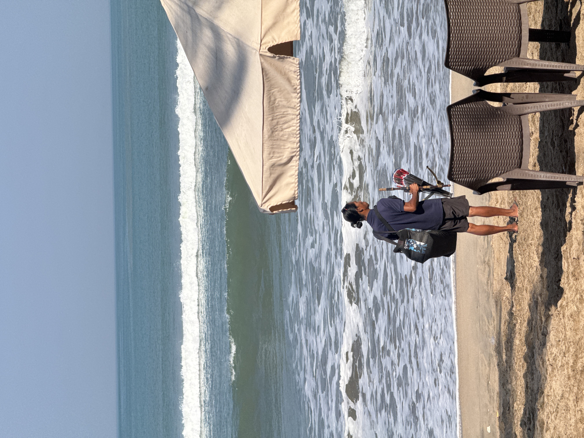 A man stands on a sandy beach in Kuta, Indonesia, facing the ocean. He has dark hair pulled back in a bun and is wearing a dark blue polo shirt and black shorts. He is carrying a large black bag over his left shoulder and holding a crossbow-like object with a fan of red and white feathers attached. The ocean is blue with white waves crashing on the shore, and the sky is clear. In the foreground, two empty brown plastic chairs are set up on the sand, and a large, light-colored beach umbrella partially shades the scene from the right. The sand is light brown and shows textures from the tide and shadows from unseen objects.