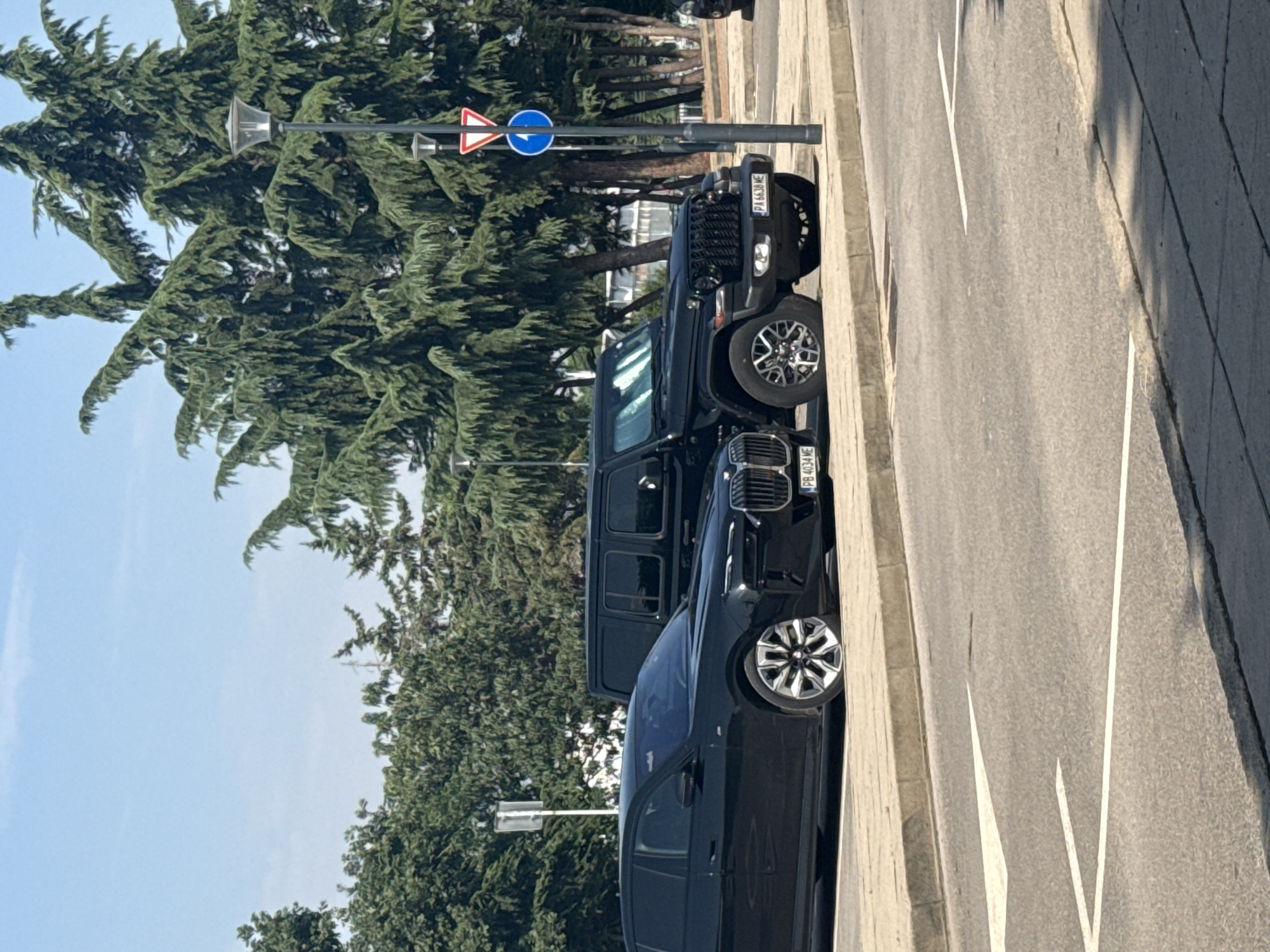 The image shows two black cars parked side-by-side on a street in Sofia, Bulgaria. The car on the left appears to be a BMW i7, and the car on the right is a black Jeep Wrangler. Both cars are facing the viewer. The BMW has a license plate that reads "PB 4034 ME". The Jeep has a license plate that reads "PA 6638 ME". In the background, there are tall green trees, a lamppost, and a blue circular traffic sign with a white arrow pointing right. The street is paved, with white lane markings visible. The sky is clear and blue.