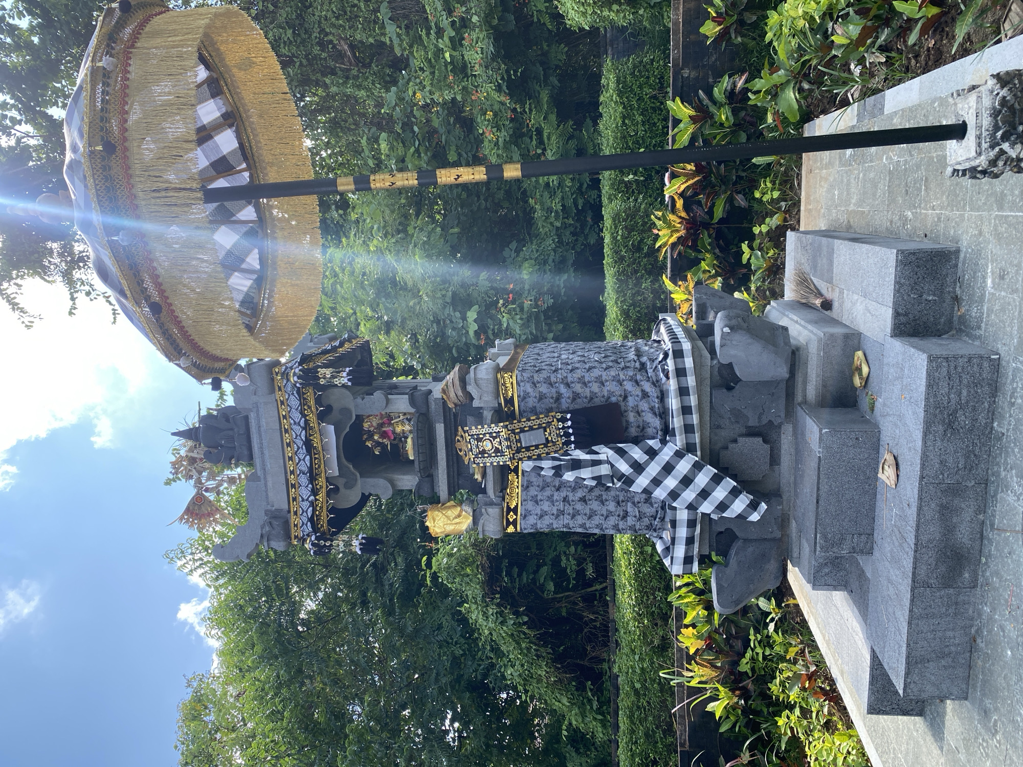 A stone shrine or temple, adorned with a black and white checkered cloth and a decorative umbrella, stands in Kuta Selatan, Indonesia. The shrine is constructed from grey stone blocks, with intricate carvings and ornaments. A traditional Balinese umbrella, featuring a gold-fringed canopy and a black and gold striped pole, provides shade. Lush green foliage, including trees and bushes, forms the backdrop, with a glimpse of blue sky and clouds visible in the upper left corner. The ground surrounding the shrine is covered with plants and foliage.