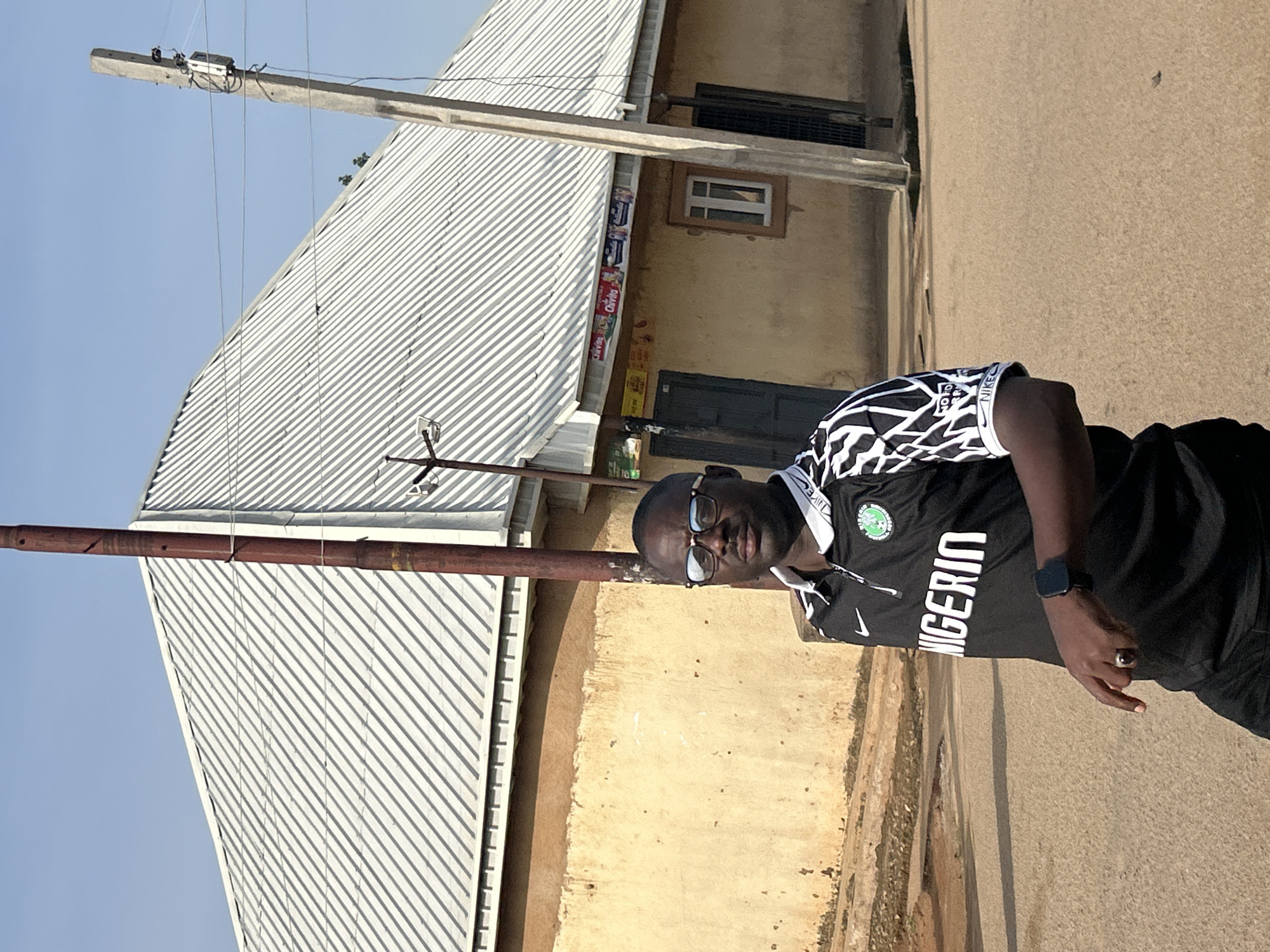 A man is standing in front of a yellow building with a metal roof. He is wearing a black jersey with "NIGERIA" written in white letters across the front. He has glasses on and is wearing a watch on his left wrist. There are utility poles and wires in the background. The ground is paved.