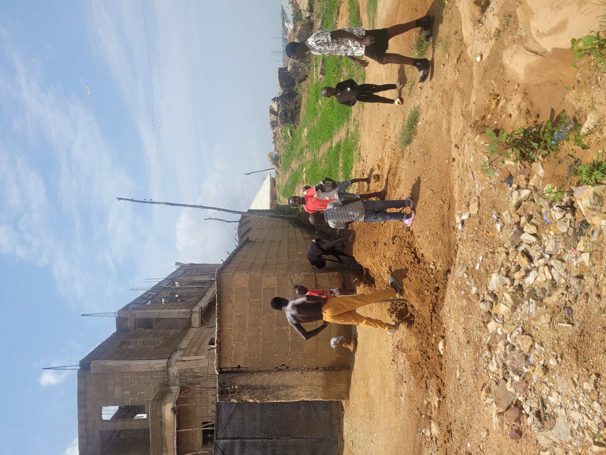 In Jos, Nigeria, under a partly cloudy sky, a group of children are engaged in various activities near a construction site. In the foreground, a shirtless young man with dark skin wears yellow pants and is seen standing with his hands on his hips, holding a shovel. Next to him, a pile of excavated soil suggests ongoing work. To his right, a younger child in a red shirt and blue shorts stands watching. Further back, another child in a plaid shirt and jeans is also present. To the far right, a young man in a white t-shirt with a patterned design and black shorts walks away from the viewer, while a smaller child in black attire stands nearby. Behind the children, a partially constructed building with cinder block walls and exposed rebar dominates the background. A large metal gate is visible to the left of the construction. The ground is a mix of red earth and gravel, with some scattered rocks and debris. The overall scene depicts a casual, community-oriented environment where children are present amidst construction activities.