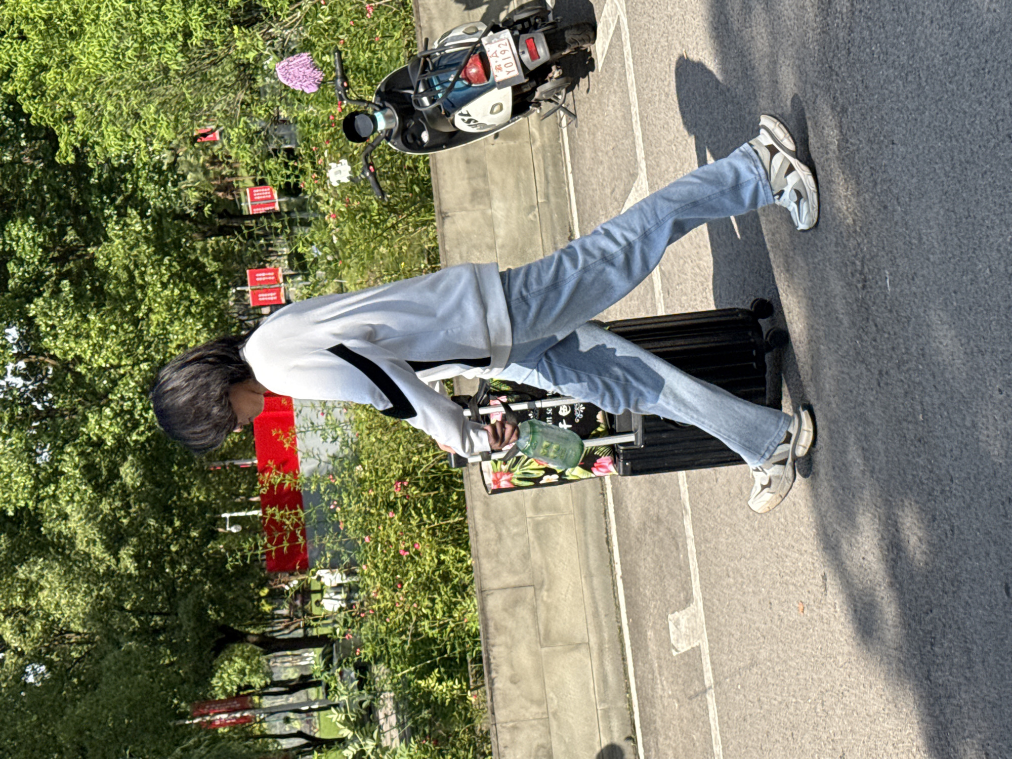 The image, taken in Jiangjin, China, shows a person walking outdoors on a sunny day, pulling luggage. The individual, appearing to be female with long dark hair, is dressed in a light-colored long-sleeved top with black accents, light blue jeans, and white, grey, and black chunky sneakers. She is pulling a black hard-shell suitcase, on top of which rests a fabric bag with a floral design, revealing partial text like "um se". In her right hand, she holds a green water bottle.
The setting is a paved path or road with clear white markings, bordered by lush green foliage, trees, and bushes, some bearing small pink and red flowers. Several red rectangular signs, with unreadable white text, are visible among the greenery. Parked on the paved surface in the background is a light-colored scooter or motorcycle, displaying the license plate "渝 A YD192". The strong sunlight casts distinct shadows on the ground, indicating a clear, bright day. The scene suggests a person in transit or on an outing.