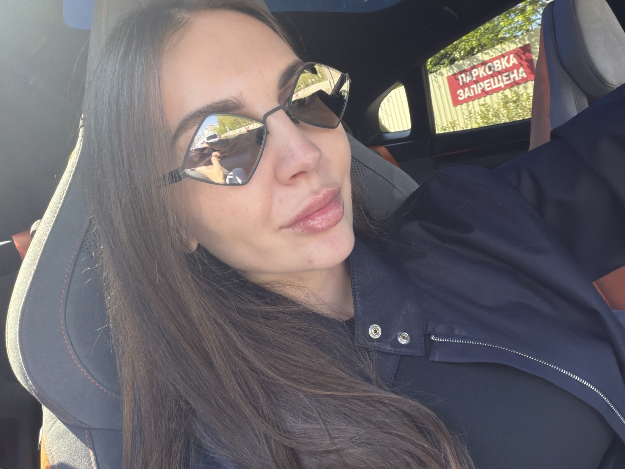 A woman with long brown hair is sitting in the driver's seat of a car. She is wearing sunglasses and a black jacket. A sign reading "ПАРКОВКА ЗАПРЕЩЕНА" (Parking Prohibited) is visible through the car window. The location is Krasnoyarsk, Russia.