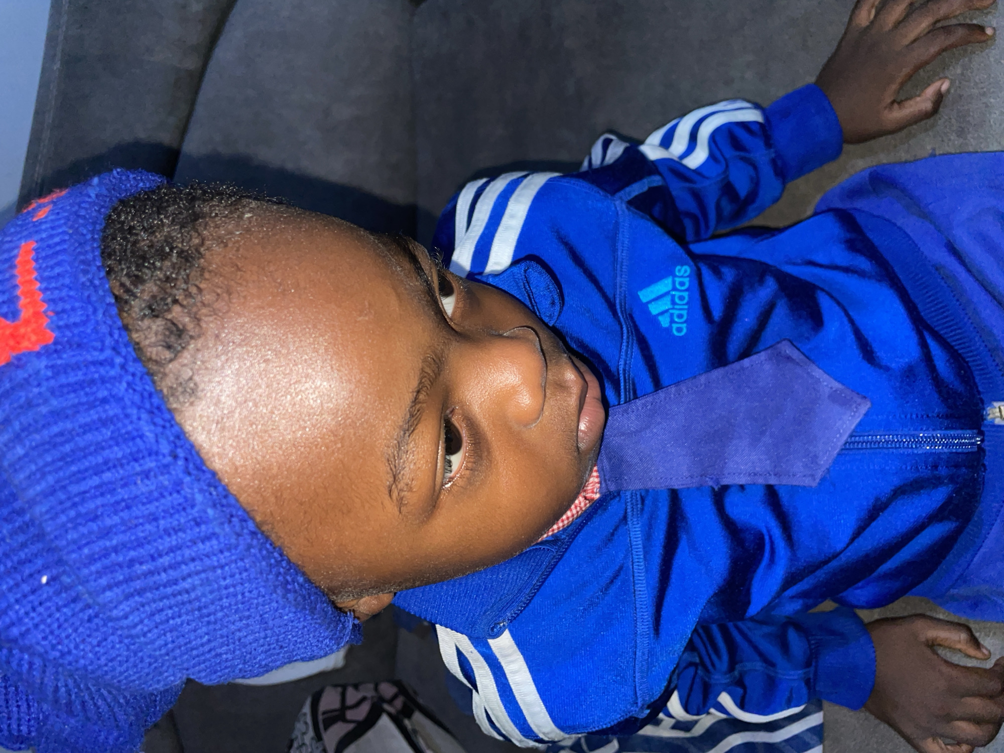 A child lies supine on a gray upholstered surface. The child wears a blue knit cap with a red design, a blue Adidas tracksuit with white stripes on the sleeves, and a blue necktie. The photograph was taken indoors.
The location in the city of Jos, Nigeria is unknown.