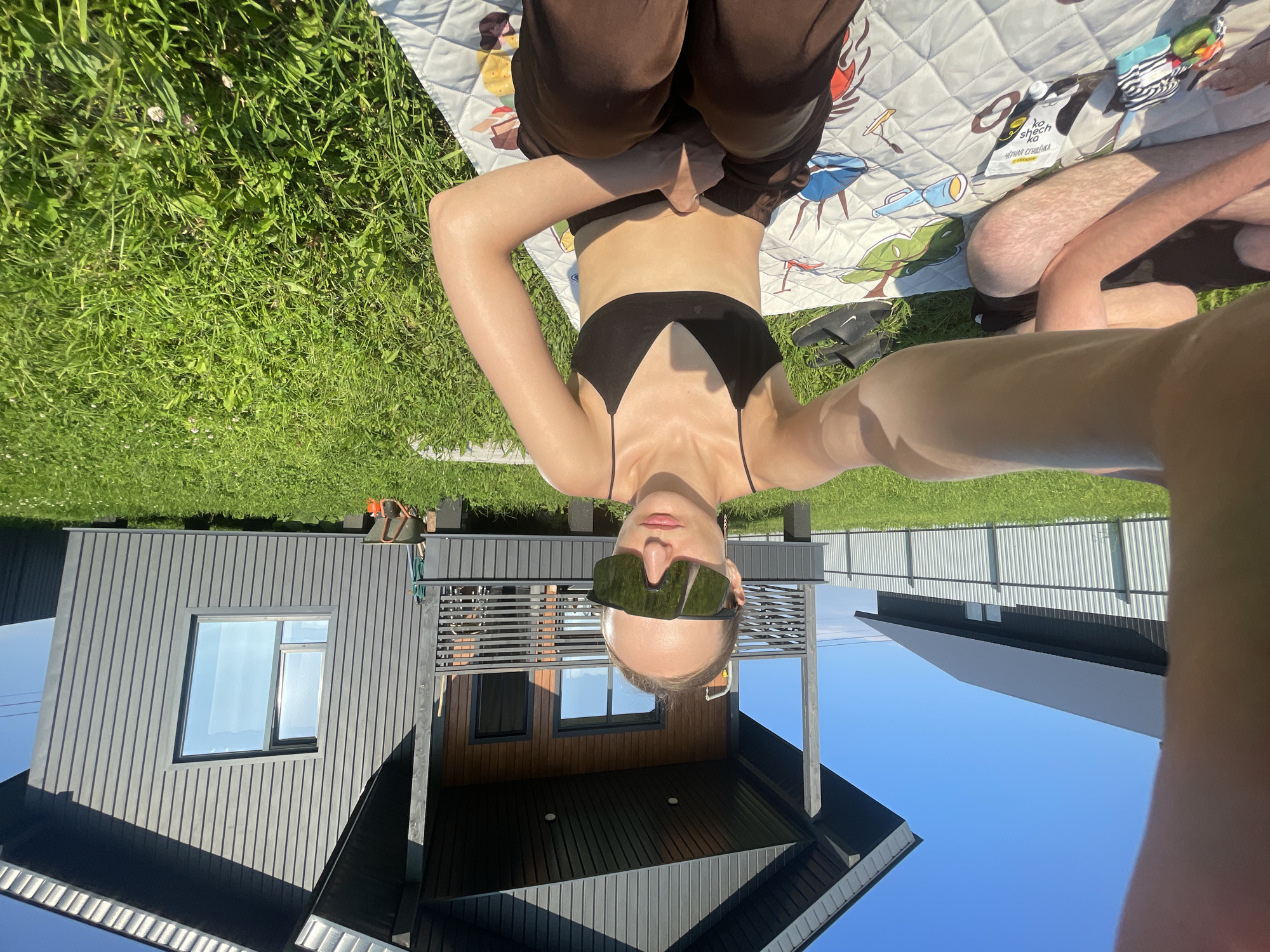 A person is lying on a blanket in a grassy yard. The person is wearing sunglasses, a black bikini top, and brown pants. A house with dark siding and windows is visible behind the person. The sky is clear and blue, suggesting a sunny day. The image appears to be a selfie, taken from a low angle. There is a faint reflection in the sunglasses, but it is unclear what it is. There is a sign in Russian on the blanket that reads "кошачий дом" which translates to "cat house". The location is given as Podshivalovo, Russia.