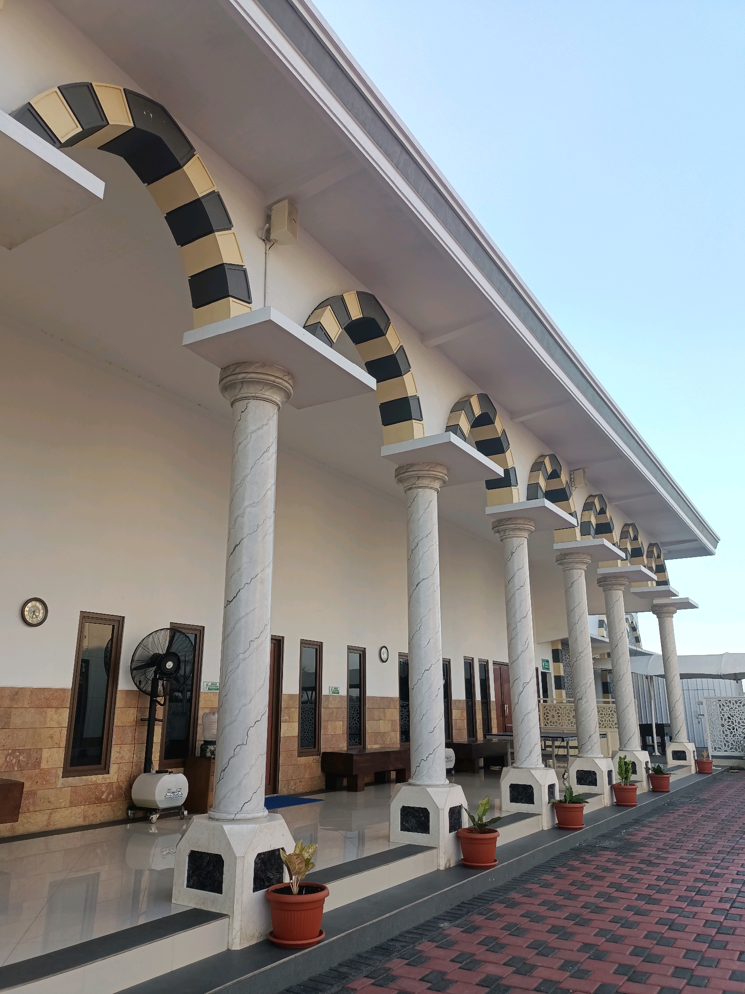 The image shows the covered walkway of a building, likely a mosque, in Plemahan, Indonesia. A series of white marble-patterned columns support arches decorated with alternating black and gold squares. The walkway has a tiled floor and a white wall behind it, with several windows and a black standing fan. Potted plants are placed in front of the columns. The sky is clear and blue. The architectural style suggests a modern interpretation of traditional Islamic design.