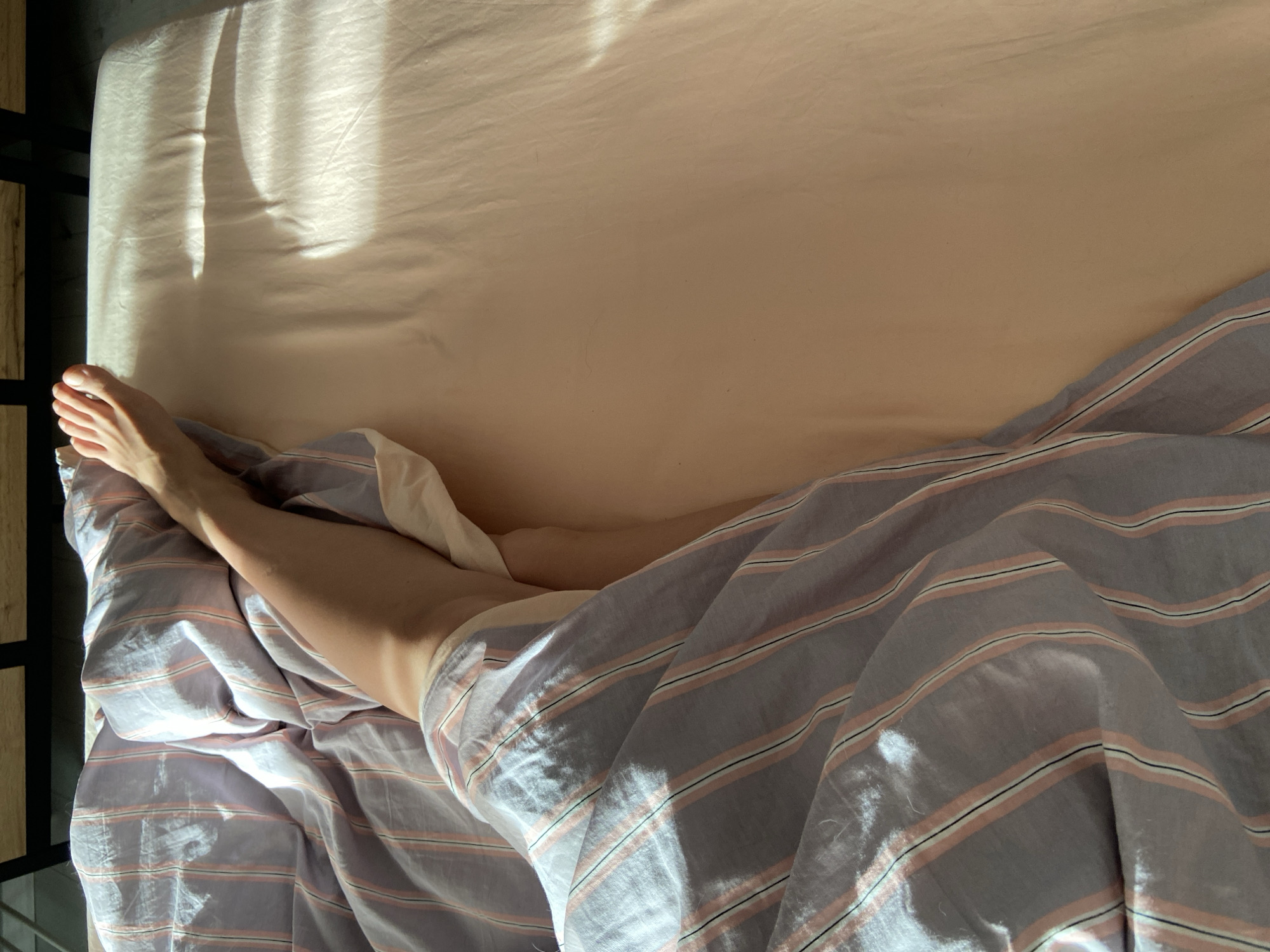 An overhead shot shows a person's bare leg and foot emerging from under a striped blanket on a bed. The blanket has vertical stripes of grey, pink, and black. The bedsheets are a pale peach color. Sunlight is streaming in from the right, creating bright highlights and shadows on the bedsheets. The scene appears to be in a bedroom.