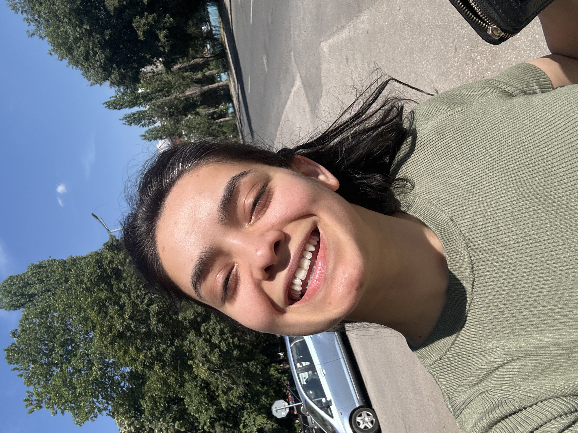 The image is a selfie taken outdoors, likely during the daytime under sunny weather. The main subject is a young person with dark hair, wearing a green ribbed top. They are smiling broadly with their eyes closed, conveying a sense of joy or happiness. Part of a black purse or wallet with a zipper is visible in the upper right corner.
In the background, a paved road or sidewalk is visible, along with a silver car parked on the side. Large, leafy green trees line the background, suggesting a park or a tree-lined street. The sky is a clear blue with a few wispy clouds, indicating a pleasant day. The overall setting appears to be a public outdoor space. There is no visible text in the image. The photo's orientation is rotated.