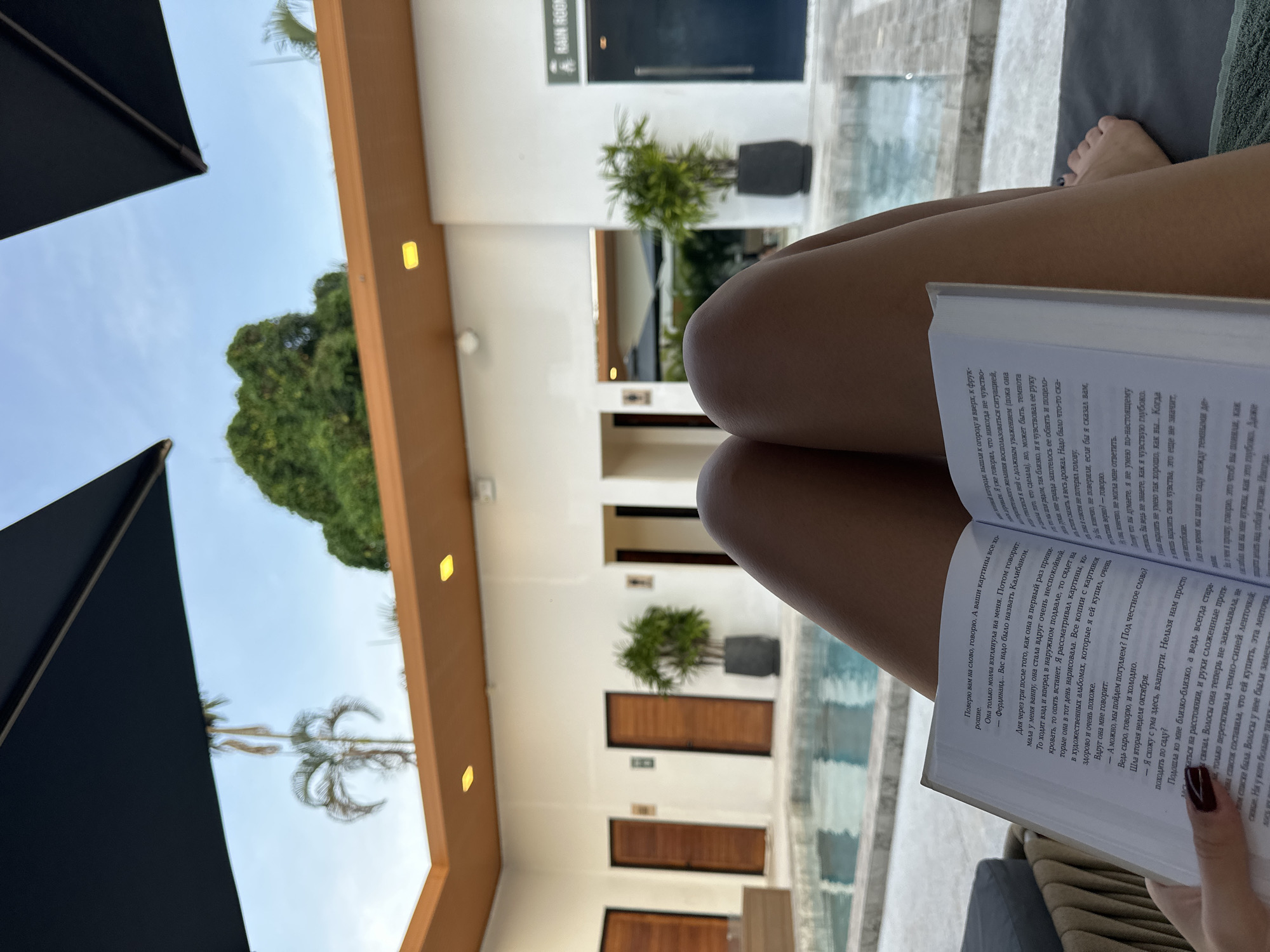 In Si Sunthon, Thailand, a person reclines on a dark grey lounge chair, holding an open book and reading. The book's pages display Russian text. The person's legs and bare feet are visible, with one foot positioned near the edge of the chair. In the background, a swimming pool reflects the sky and surrounding architecture. The area is characterized by white walls, wooden doors, potted plants, and overhead awnings. The scene appears to be an outdoor patio area, possibly part of a resort or private residence, with a tropical ambiance suggested by the palm trees visible in the distance. The lighting indicates it is daytime.