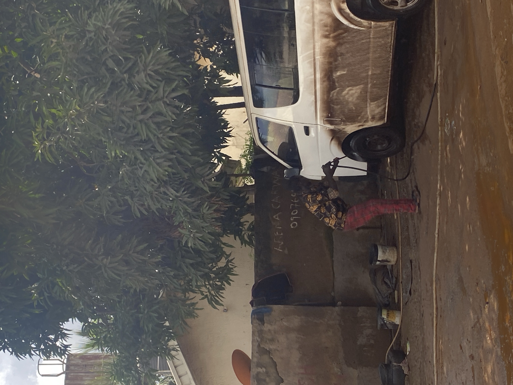In Katambe, Nigeria, a person is washing a white van at an outdoor car wash. The van is heavily soiled with mud, particularly on its lower half. The person, wearing a patterned shirt and red and black plaid pants, is using a pressure washer to clean the vehicle. To the left of the van, there are several buckets on the muddy ground. Behind the person and to the left, a wall with writing on it is visible, reading "ARENA CARWA" and "090769". A large tree with dense green foliage overhangs the scene. The environment appears to be a utilitarian outdoor space, with muddy water and scattered cleaning supplies.