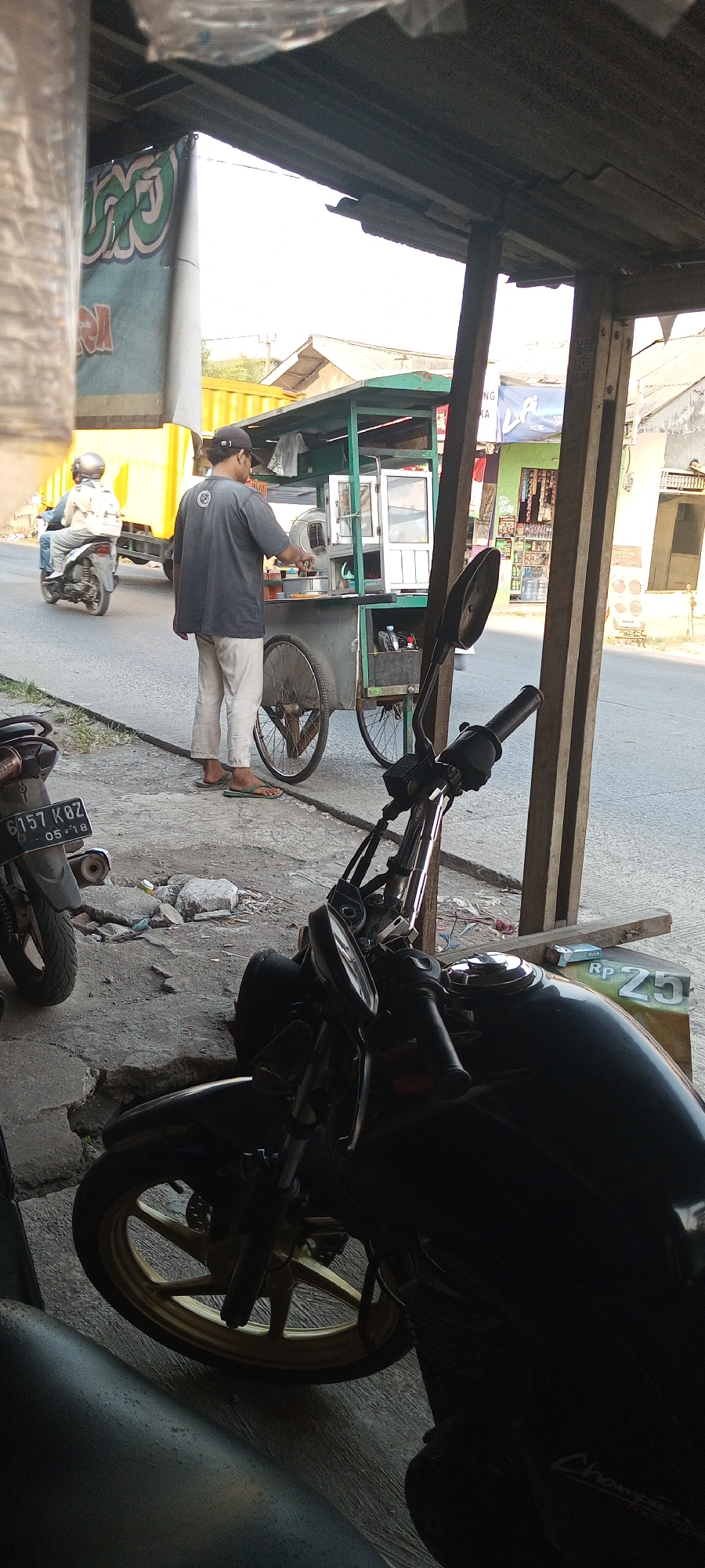 In Setu, Indonesia, a man wearing a dark grey t-shirt and light-colored pants stands by a food cart. He is facing the cart, with his right hand extended and holding what appears to be a utensil. The food cart is green and grey, with wheels and a structure for displaying food. Behind him, a yellow truck is visible, and further back, buildings and houses line a street. To the left of the man, a person wearing a helmet is riding a motorcycle. In the foreground, a black motorcycle is parked, with its front wheel and handlebars visible. The scene appears to be outdoors, with natural light suggesting daytime.