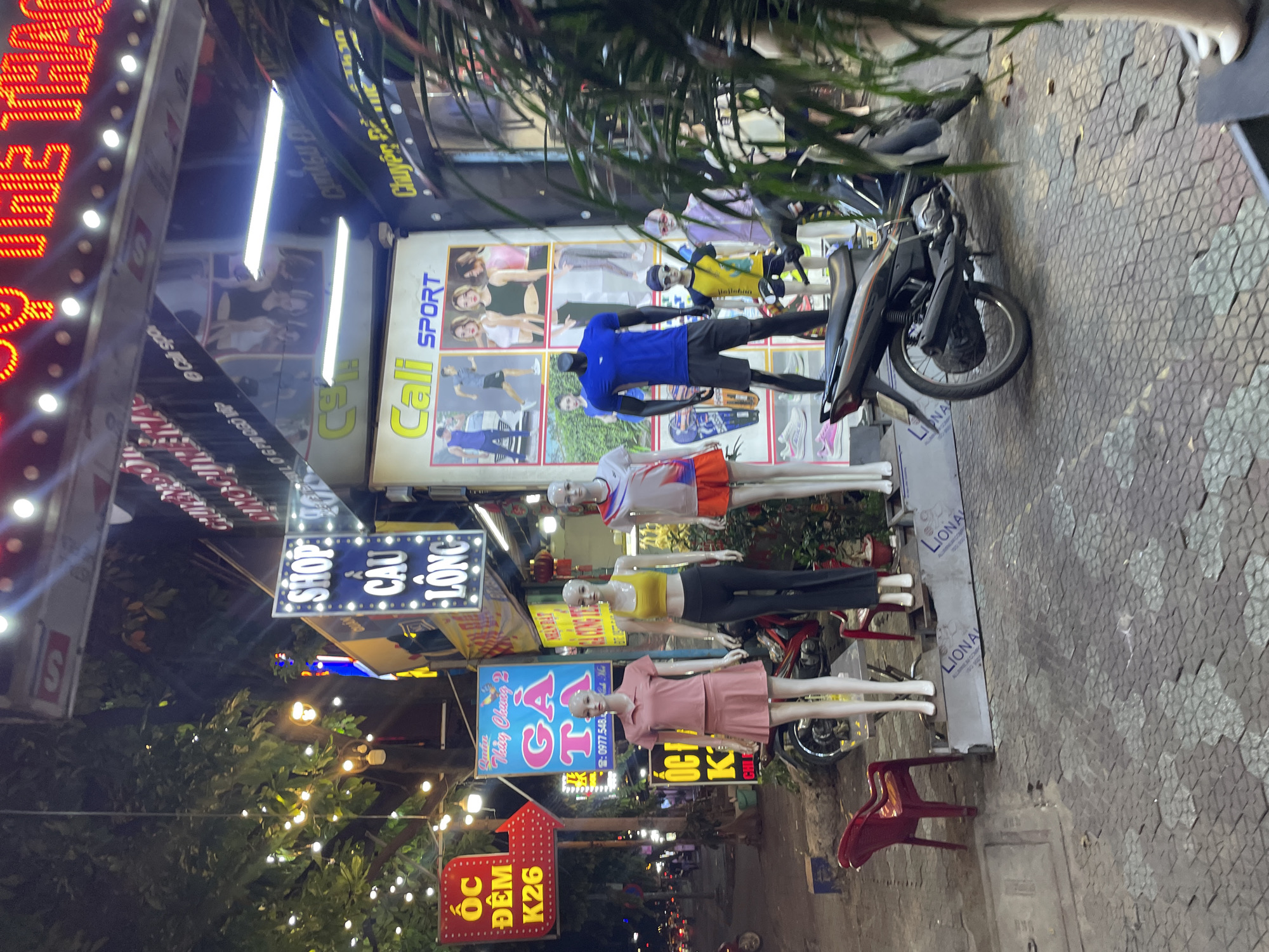This is a night time, eye-level, medium shot of a street-level storefront in Thuận An, Vietnam, likely a clothing store specializing in sportswear. The scene is illuminated by bright neon signs and artificial lighting, creating a vibrant atmosphere.
In the foreground, several mannequins are displayed. One female mannequin on the left wears a pink polo shirt and skirt set. Next to her, another female mannequin is dressed in a yellow sports bra and black leggings. A third female mannequin on the right is clad in a white and red t-shirt with orange shorts. A male mannequin stands to the left of the yellow-bra-clad mannequin, wearing a blue t-shirt and dark shorts. A child mannequin, wearing a yellow and blue t-shirt and shorts, stands in front of the male mannequin. To the right of the child mannequin, another mannequin is partially visible, wearing a light purple outfit. The mannequins are displayed on a raised platform in front of the store.
Behind the mannequins, there are several signs. A prominent blue and white sign reads "SHOP" in large letters at the top, followed by "CÂU LÔNG" below, which translates to "Badminton" in Vietnamese. Another sign to the left is red with white lettering and an arrow, reading "ỐC ĐÊM K26" which likely indicates a late-night snail dish. A large poster behind the mannequins features images of people in athletic wear, with the text "Cali SPORT" visible.
A motorcycle is parked in front of the store, partially obscuring the lower part of the mannequins. A red plastic chair is placed on the sidewalk to the left of the mannequins. The ground is paved with hexagonal tiles. In the background, trees with decorative lights are visible, along with other storefronts and street activity, including traffic and pedestrian lights. The overall impression is a lively commercial street scene at night.