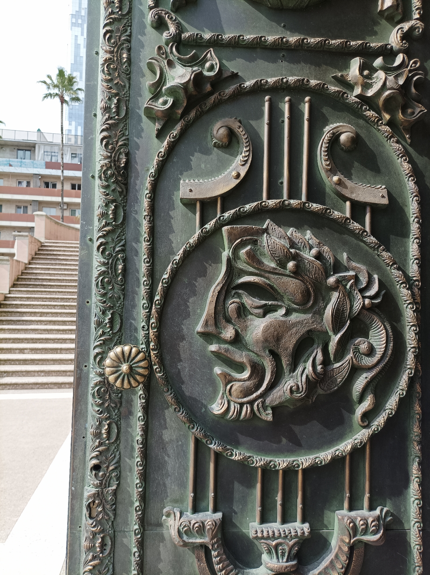The image depicts a close-up of an ornate, likely bronze, door panel in Batumi, Georgia. The panel features a raised relief sculpture of a mythological figure's head within a circular frame. Ornate, curvilinear designs and vertical bars are incorporated into the door's design. A decorative door handle with a floral pattern is visible on the left side of the panel. The door's surrounding frame is heavily ornamented. In the background, a stairway ascends to a building, and a palm tree rises behind the building. A modern building is also partially visible in the far background. The scene is bathed in sunlight, creating shadows that highlight the intricate details of the door panel.
