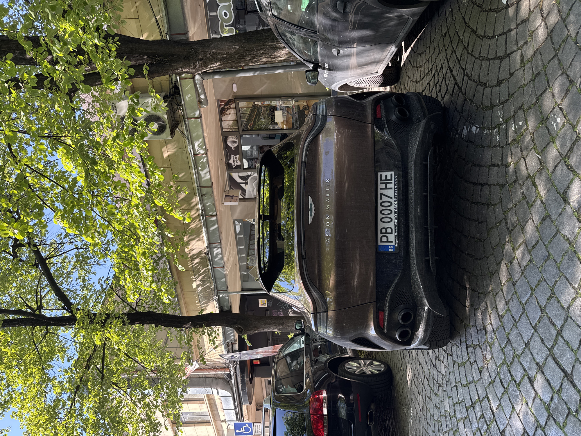 The image depicts the rear of a brown Aston Martin parked on a cobblestone street in Plovdiv, Bulgaria. The license plate reads "PB 0007 HE". Other vehicles are parked around the Aston Martin. The buildings in the background include businesses with signage, and a tree with green leaves is present on the left side of the frame. An air conditioning unit is visible on the building. A handicap parking sign is also seen near a dark-colored vehicle. The sky is visible through the tree's foliage.
