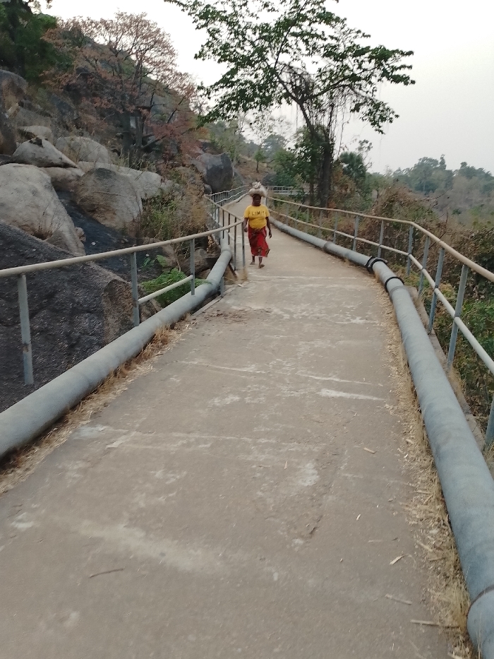 A woman in a yellow t-shirt and red skirt walks along a concrete path lined with metal railings and large pipes. She carries something on her head. The path runs alongside a rocky, tree-covered hillside on the left and greenery on the right. The background suggests a natural, elevated landscape. The sky appears hazy or overcast.