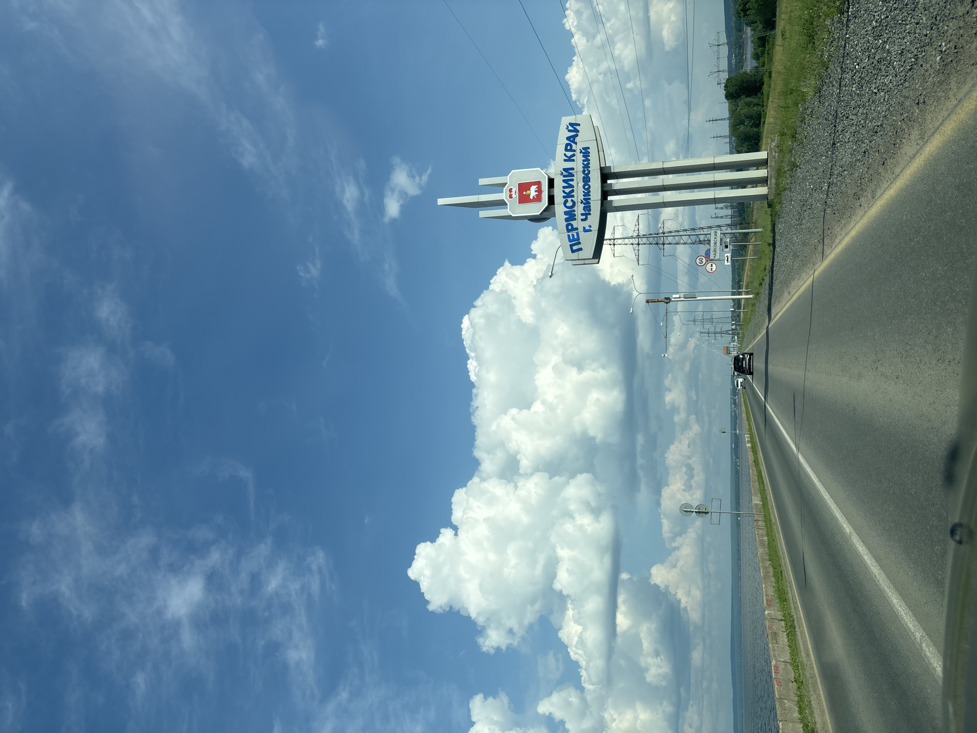 The image shows a road leading towards a large sign that reads "ПЕРМСКИЙ КРАЙ г. ЧАЙКОВСКИЙ" (Permskiy Krai, City of Chaykovsky) in Russian. The sign is white with blue lettering and has a red emblem at the top. The road is asphalt with white lane markings and a yellow edge line. To the left of the road is a large body of water, possibly a river or lake, with a concrete barrier along its edge. To the right of the road are power lines, tall metal towers, and a grassy area with trees in the distance. The sky is blue with scattered white clouds. Several cars are visible on the road ahead. The perspective suggests the photo was taken from inside a vehicle. The sign indicates the location is the border of Perm Krai and the city of Chaykovsky in Russia.
