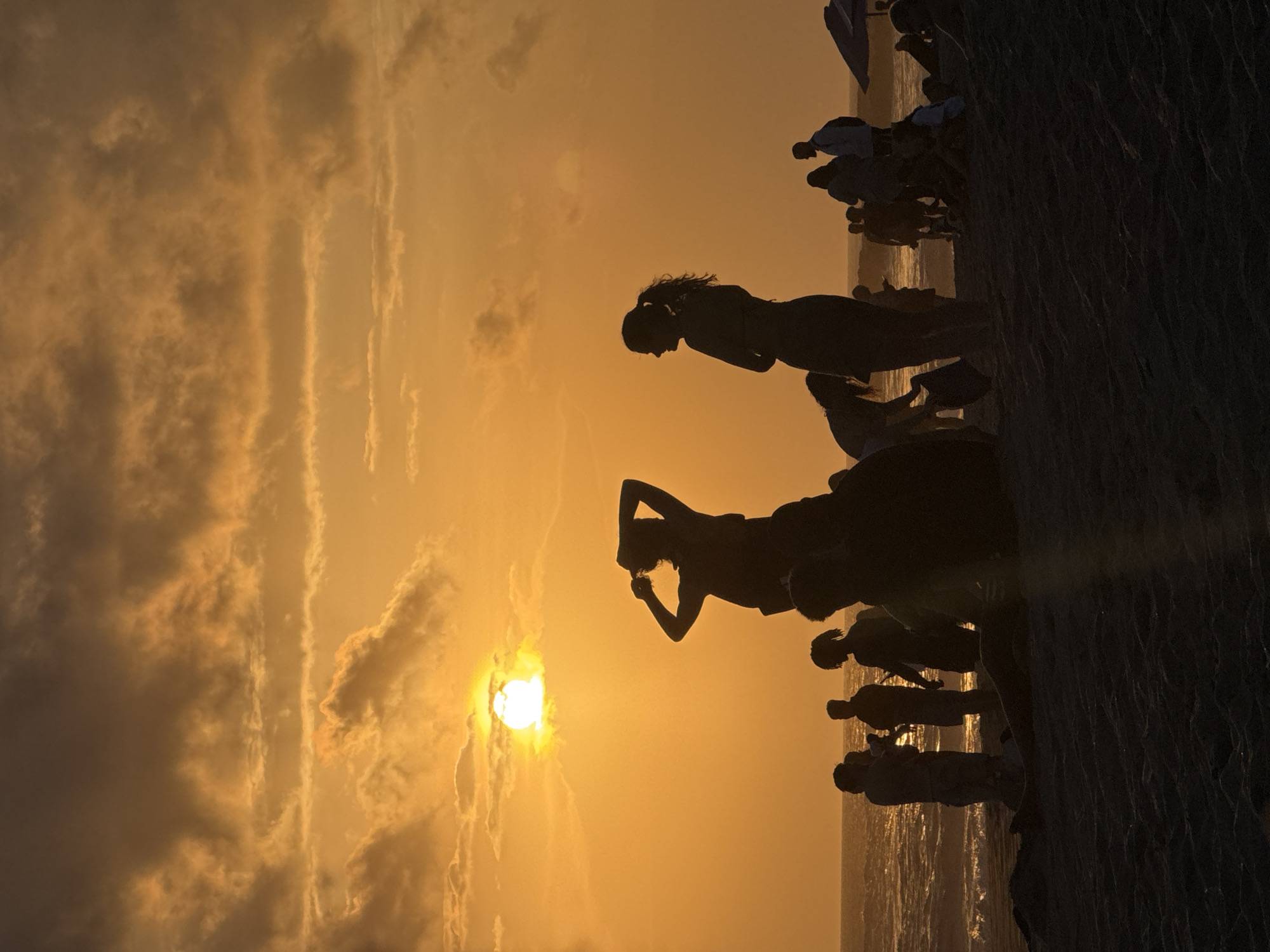 The image shows a silhouette of people on the beach against a sunset. The location is North Kuta, Indonesia, and the people are gathered to watch the sunset. The sun is low on the horizon and casting a golden light on the clouds and the water. Some people stand and pose for pictures. Some of the people are partially obstructed by dark vegetation on the far right of the picture.