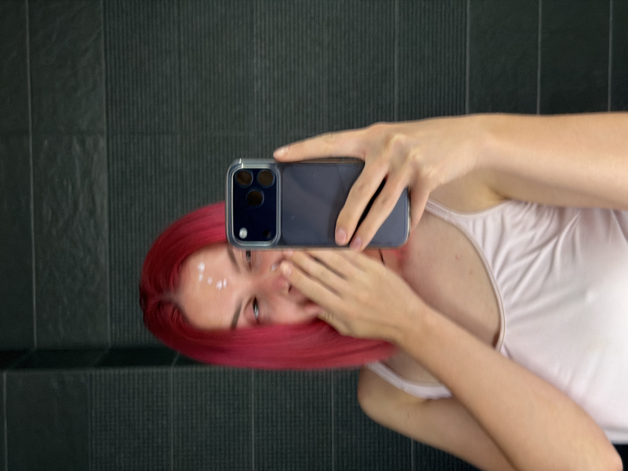 A young person with bright pink hair is taking a selfie in a mirror. They are wearing a white tank top and have white dots of cream on their forehead. Their right hand is covering their mouth as they look into the phone. The background appears to be a tiled wall with horizontal lines, possibly in a bathroom. The image does not contain any information that would indicate the location is in Chaikovsky, Russia.