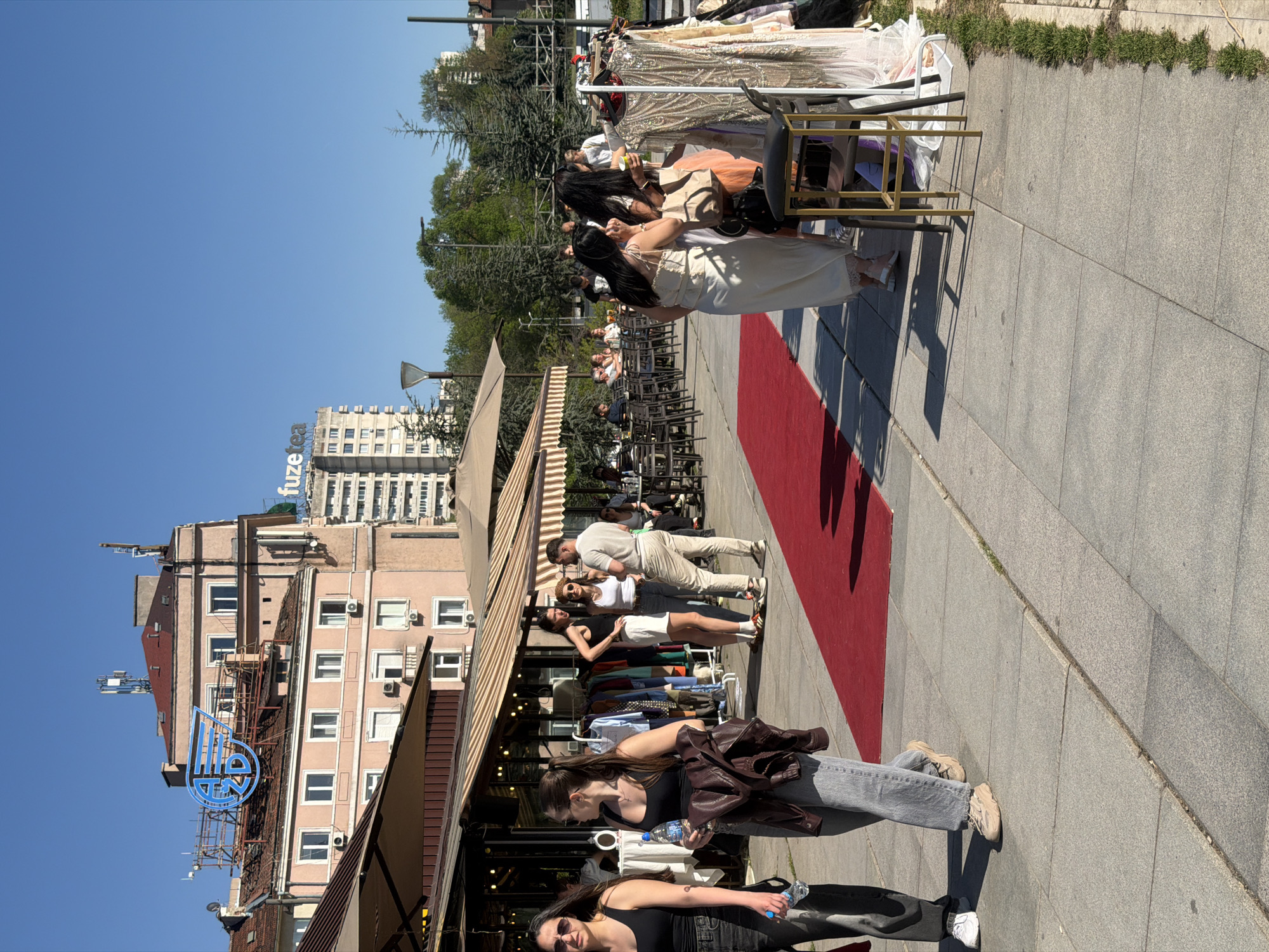 The image shows a street scene in Sofia, Bulgaria. Several people are walking on a wide sidewalk. Some are looking at clothing displayed on racks in front of a store under a striped awning. A red carpet runs down the middle of the sidewalk. Several people appear to be posing in front of more clothing displayed at the end of the carpet. Buildings are in the background, including one with a large "fuze tea" sign on top and another with a blue logo. The sky is clear and blue.