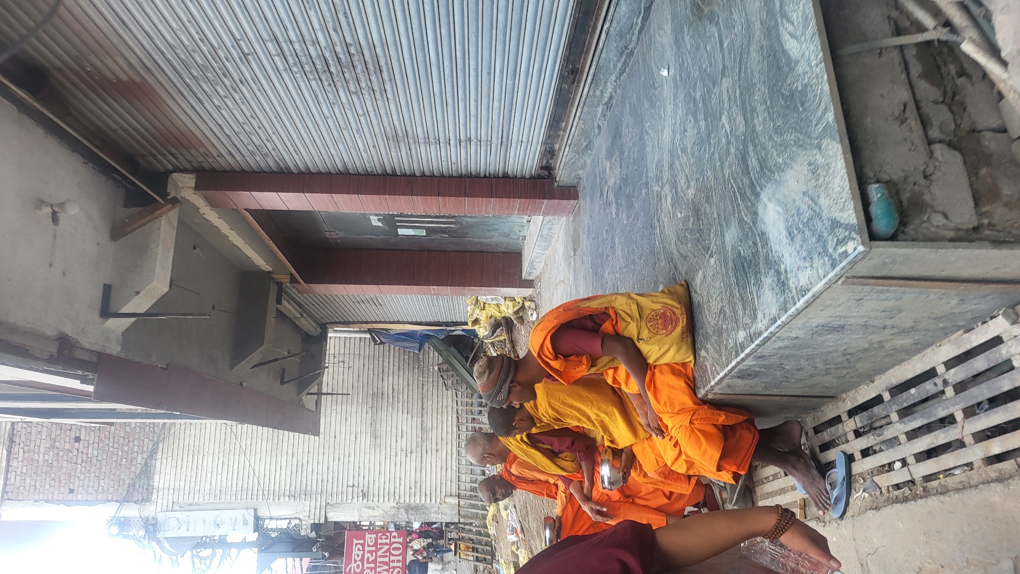 Four Buddhist monks sit along a concrete curb in Dharamshala, India. They wear orange robes. Each monk holds a small metal bowl. A blue sandal rests on the ground near the curb's edge. A blue ceramic vase sits on the curb. A metal roll-up door forms the background. Shop signs are visible in the distance.