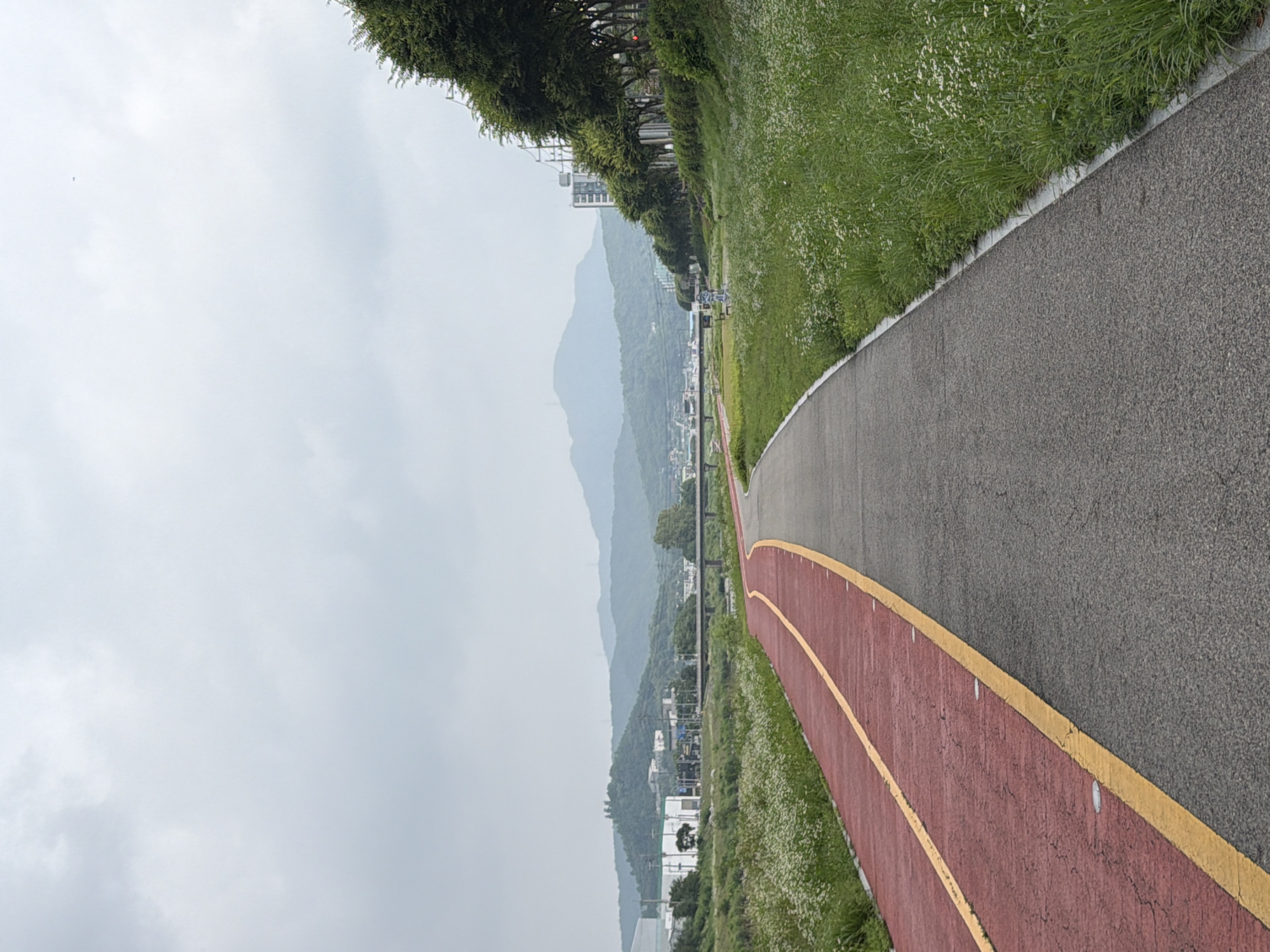 A paved walking and cycling path curves through a green landscape. The path is divided into two lanes: a red lane for cycling and a grey lane for walking. The path is bordered by lush green grass with small white flowers. In the distance, rolling hills are visible, covered in trees and vegetation. A bridge spans across a body of water, and several buildings, including some apartment complexes, are scattered among the trees. The sky is overcast with a light grey hue. The scene is located in Dongducheon-si, South Korea.