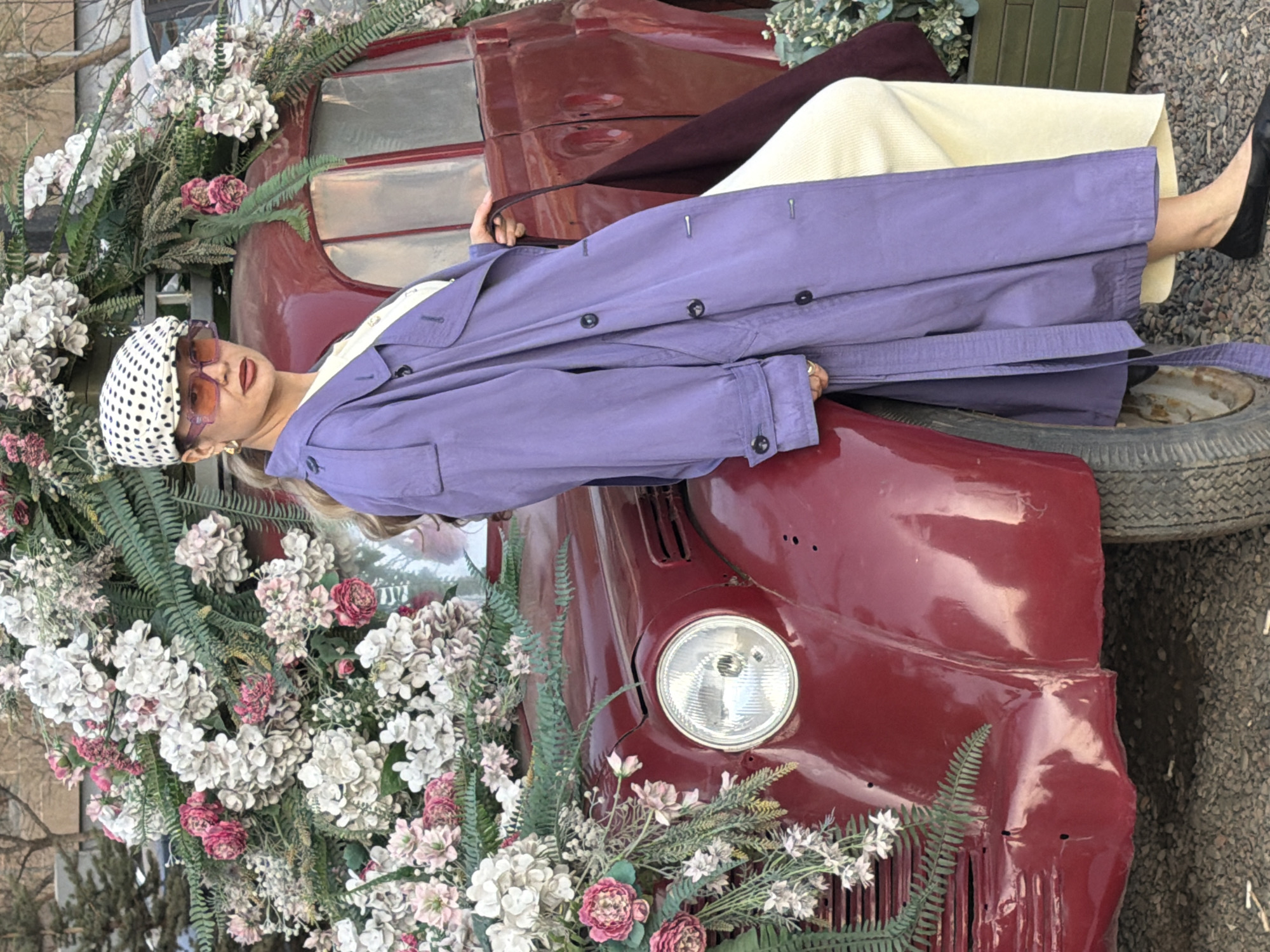 A female is reclining on a vintage maroon vehicle that is heavily decorated with white, pink, and green flowers and ferns. She is wearing a white headscarf with black polka dots, orange-tinted sunglasses, a cream-colored top and pants, a long light purple trench coat, and black shoes. The vehicle has a single headlight visible, and there is a spare tire positioned behind her feet. The ground is covered in gravel. The photo was taken in the city of Krasnoyarsk, Russia.