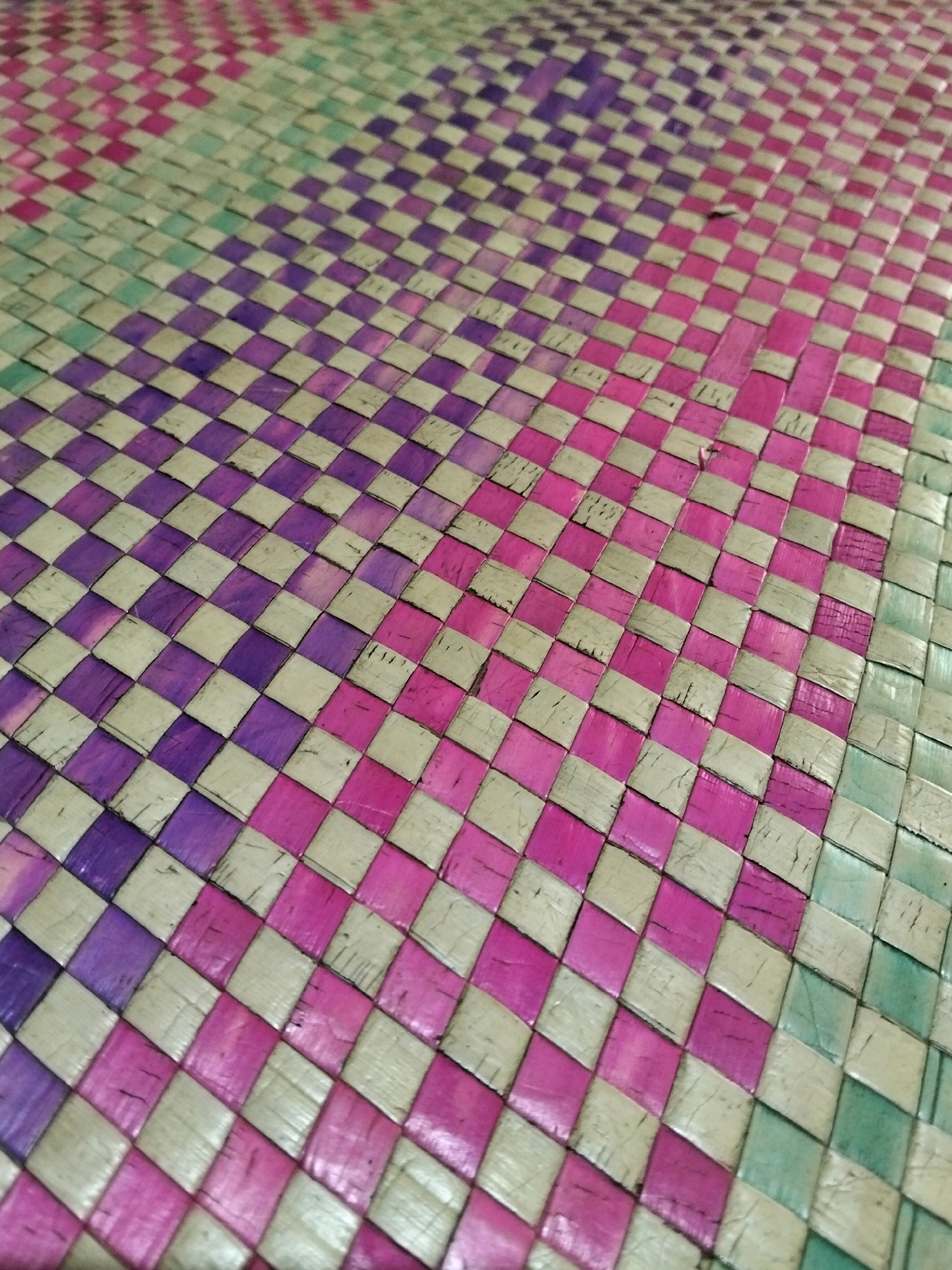 The image is a close-up, overhead shot of a woven mat with a checkered pattern. The mat is made of natural fibers that have been dyed in various colors, including pink, purple, light green, and a light beige or off-white. The weaving creates a diamond pattern where the colors alternate. The perspective is at an angle, showing the texture and depth of the woven material. The background is out of focus, highlighting the mat as the primary subject. The scene is devoid of any discernible environmental context or human presence, and there is no indication of its location within Teluk Pambang, Indonesia.