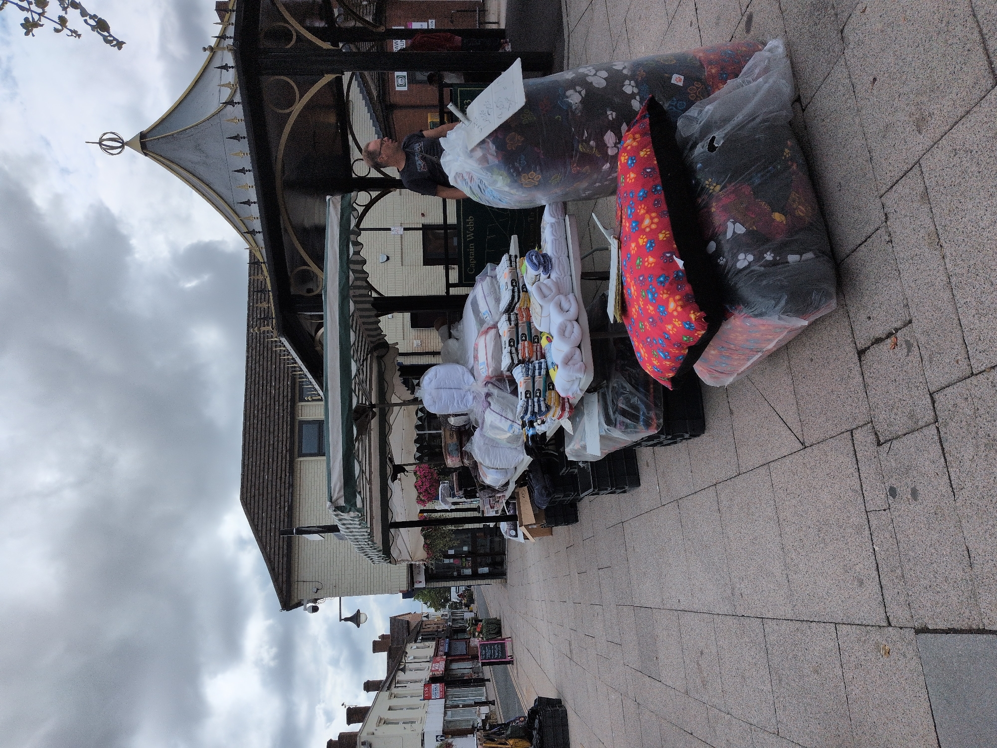 The image depicts an outdoor market stall located on a paved street in Telford, United Kingdom. The stall is laden with various items, including stacks of folded towels, bedding, and large, clear plastic bags filled with what appear to be soft goods, possibly pet beds, as indicated by paw print patterns on some of the items. A man wearing glasses and a dark t-shirt stands behind the stall, looking towards the left. In the background, a street lined with shops and buildings extends into the distance, under a cloudy sky. The architecture suggests a typical urban setting in the UK.