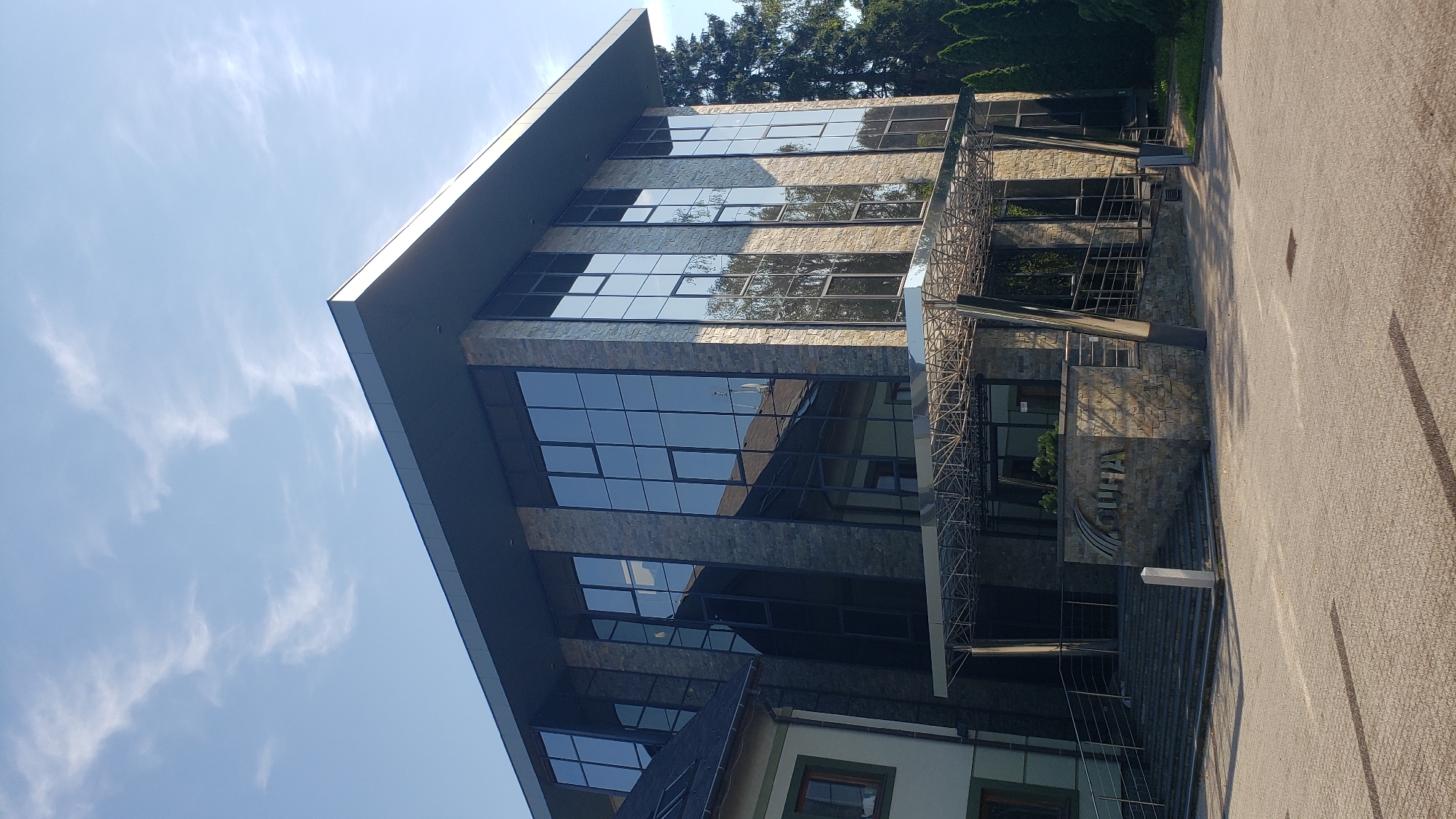 The image shows the exterior of a modern office building with a stone facade and large glass windows. A canopy with a metal frame extends over the entrance, which is accessed by a set of stairs and a ramp. A sign with the text "GUHA" is visible in front of the entrance. The building appears to be located in Prešov, Slovakia. The sky is clear with some wispy clouds. There are trees and other greenery surrounding the building. The ground in front of the building is paved with interlocking bricks. No people are visible in the image.