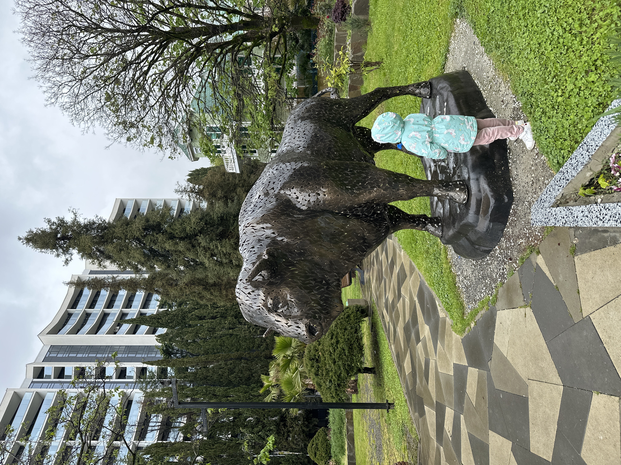 A full shot, viewed from a high angle, depicts a metal sculpture of a bull in Sochi, Russia. A child in a light blue coat and hood stands at the front of the sculpture, appearing to interact with it. The statue is situated on a dark platform surrounded by grass and a paved pathway. Behind the bull, a tree and a multi-story building with a unique architectural design are visible. Various green trees and shrubs add to the landscape. The weather seems overcast, suggesting a daytime outdoor setting.