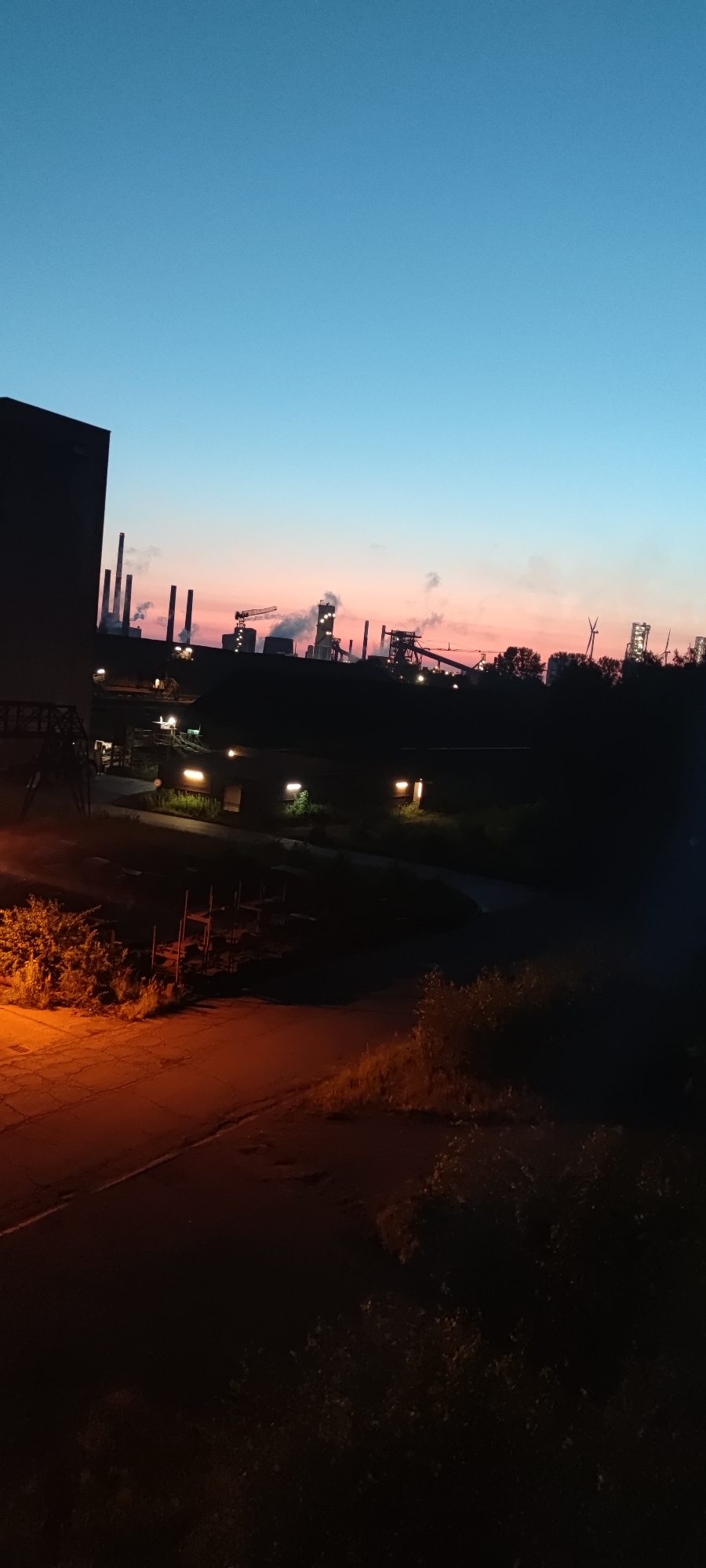 The image depicts a nighttime view of the Salzgitter-Luppersum industrial area in Salzgitter, Germany. The sky transitions from a deep blue at the top to a soft pinkish-orange hue near the horizon, suggesting either sunrise or sunset. Silhouetted against the sky are numerous industrial structures, including tall smokestacks emitting plumes of smoke, cranes, and various buildings characteristic of a steelworks or heavy industry. Lights are visible on some of the structures and in the foreground, illuminating parts of the scene. In the immediate foreground, there is a dark, paved area, possibly a road or walkway, with some vegetation bordering it. A large, dark building occupies the left side of the frame. The overall impression is one of industrial activity operating into the night, set against a twilight sky.