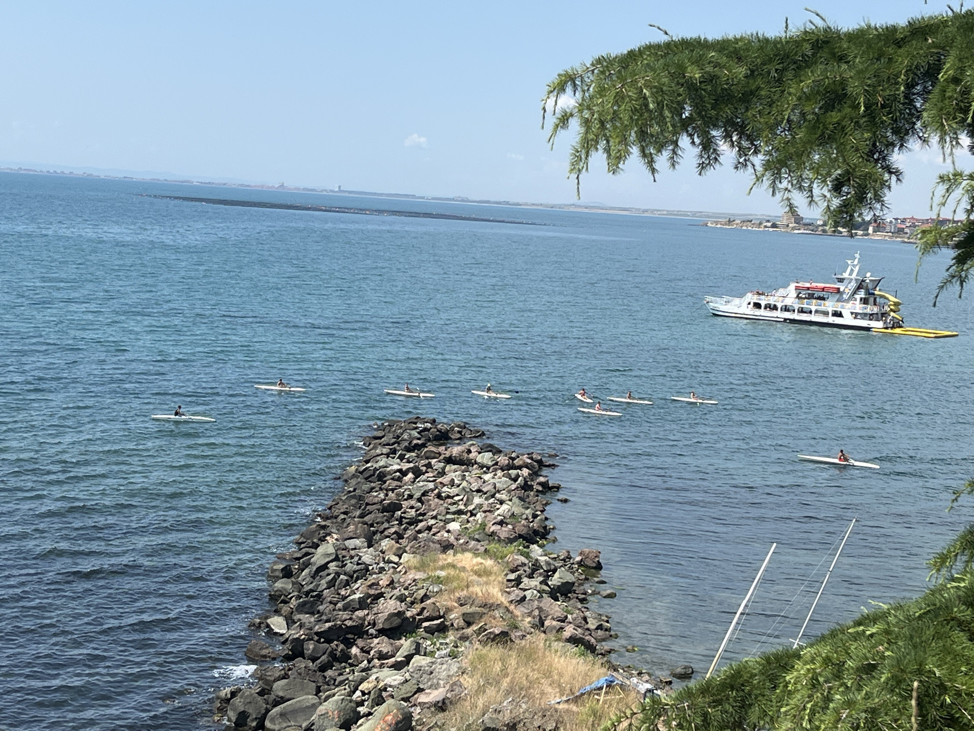 A serene daytime view captures the azure waters of the Black Sea near Nesebar, Bulgaria. In the foreground, a rocky breakwater juts into the sea, partially covered in dry grass. Further out, a group of people are kayaking, their small vessels scattered across the water. A white passenger ferry, laden with people and featuring a yellow slide, sails in the mid-ground. The distant shoreline is dotted with buildings, indicative of the historic town of Nesebar. Overhanging evergreen branches frame the upper portion of the image, adding a touch of natural beauty to the scene. Two sailboat masts are visible in the lower right corner. The sky is a clear, pale blue.