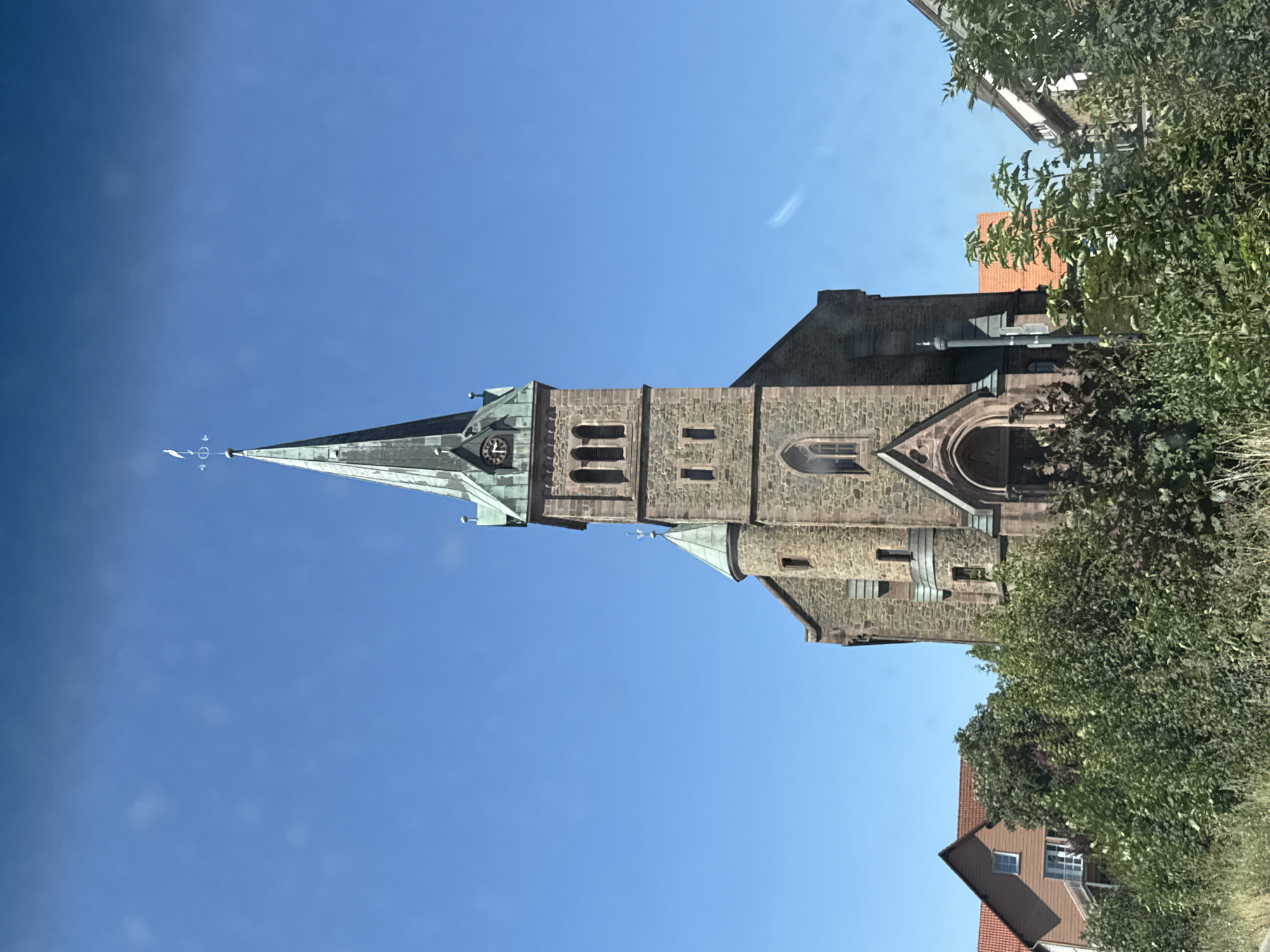 This is an outdoor, eye-level, medium shot photograph of a stone church with a tall, pointed steeple, located in Herzberg, Germany. The sky is clear and blue. The church is made of reddish-brown stone and has green copper accents on its roof and steeple. There are arched windows and doorways, and a clock face is visible on the steeple. A weather vane shaped like a bird sits atop the steeple. In front of the church, there is dense green foliage and trees. To the left of the church, a residential building with a red-tiled roof and a light brown facade is partially visible. To the right, parts of other buildings can be seen through the trees. The lighting suggests it is daytime, likely during sunny weather.