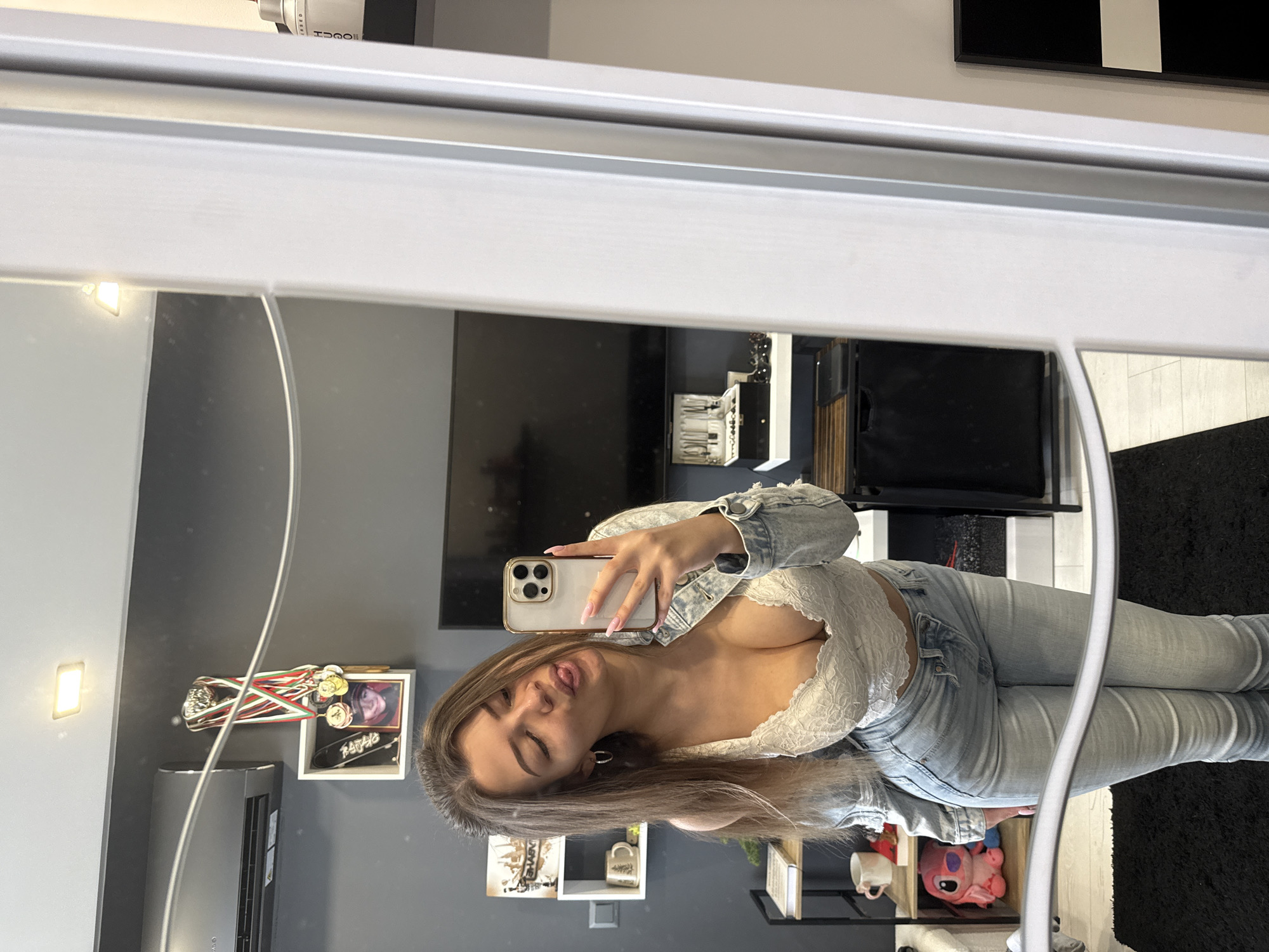 A woman is taking a selfie in a mirror. She is wearing a denim jacket, white lace top, and blue jeans. She is holding a smartphone in her right hand. Her face is partially visible, and she has long brown hair. The background includes a television, shelving unit with various items, and a framed picture. The scene appears to be indoors. The location in the city of Sofia, Bulgaria cannot be determined from the image.