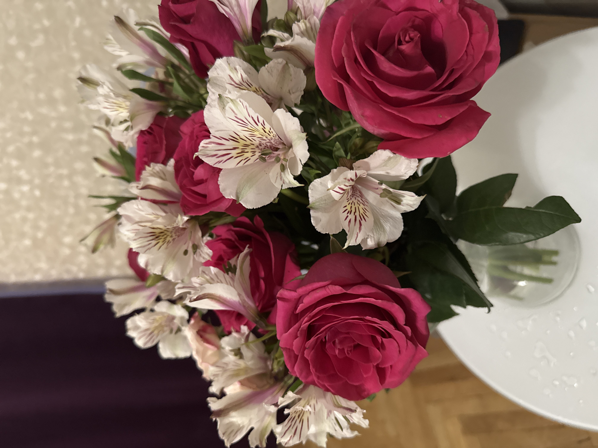 A high-angle shot depicts a bouquet of pink roses and white alstroemeria flowers in a glass vase. The arrangement sits on a white, possibly circular, table. The background shows a cream-colored wall and a glimpse of a dark surface, potentially a sofa or floor. The image suggests an indoor setting. There's no information in the image to suggest the location of Saint Petersburg, Russia.