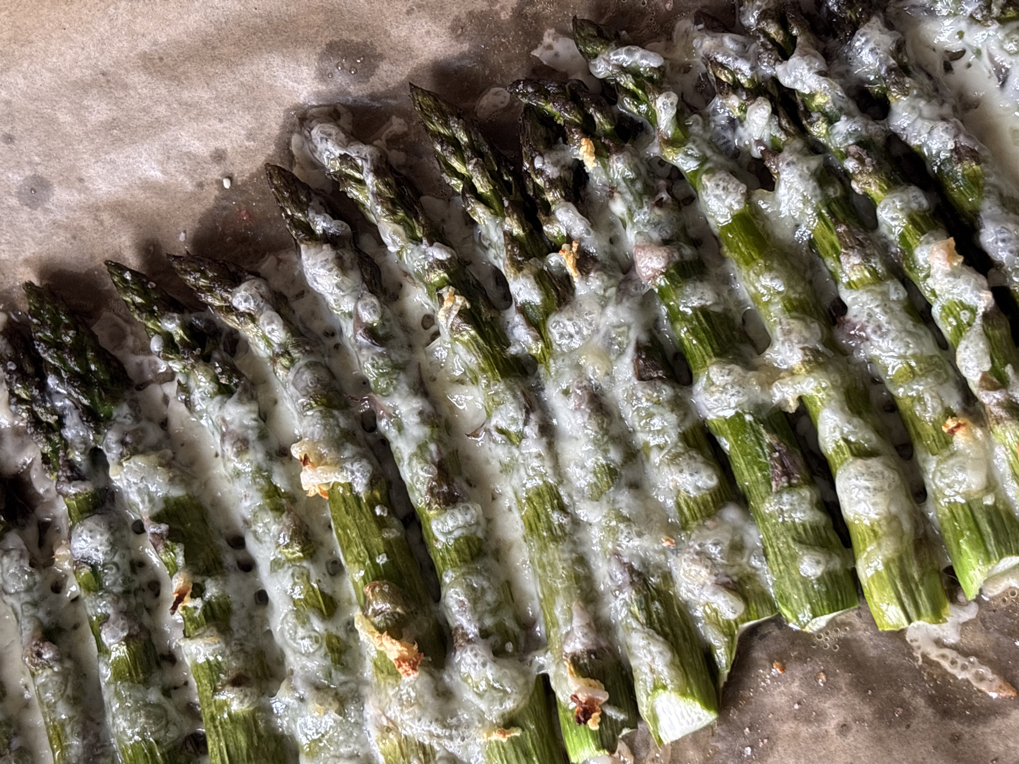 The image shows a close-up of baked asparagus on a baking sheet. The asparagus spears are arranged in a row, covered with a melted, white substance. The tips of the asparagus are slightly charred. The baking sheet is lined with parchment paper.
