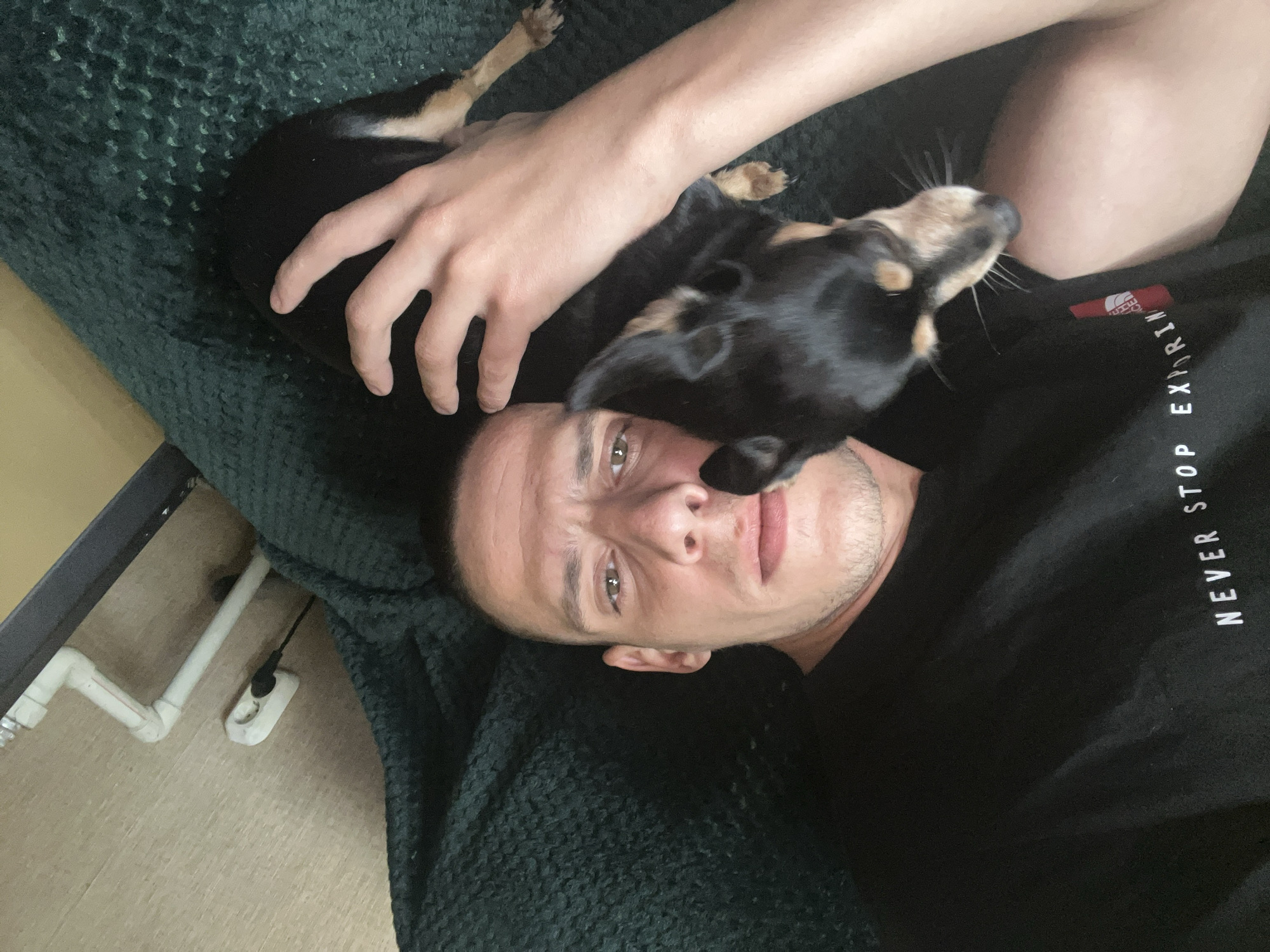 A man and a dog are lying down. The man is lying on his back, wearing a black t-shirt with white text "NEVER STOP EXPLORING". The dog is lying on top of the man. The dog is small, black and brown, with its head close to the man's face. The man has his right hand on the dog. The background is a green blanket. A white pipe is visible. The location cannot be determined from the image.