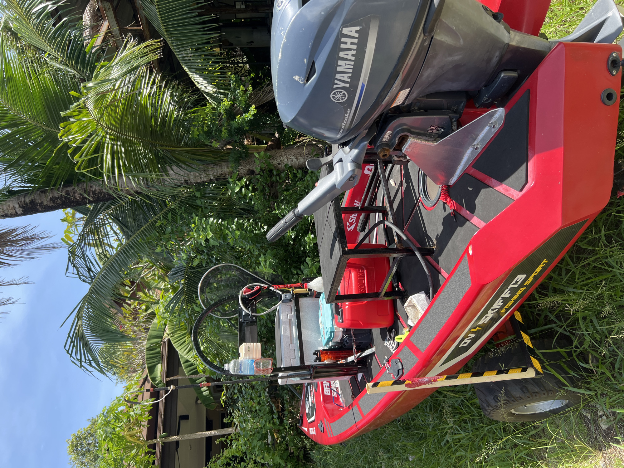 A red aluminum skiff with a Yamaha outboard motor is parked on grassy ground in front of a building with a dark roof and lush green vegetation, including palm trees, indicative of a tropical location like Choeng Thale, Thailand. The skiff is equipped with a black T-top structure supporting various items including a water bottle, a cooler, a red gas can, and fishing nets. The skiff itself displays the text "DV SKIFF 13 FISHING SPORT" along its side. The outboard motor is a Yamaha FourStroke. The environment is sunny with clear blue skies visible.